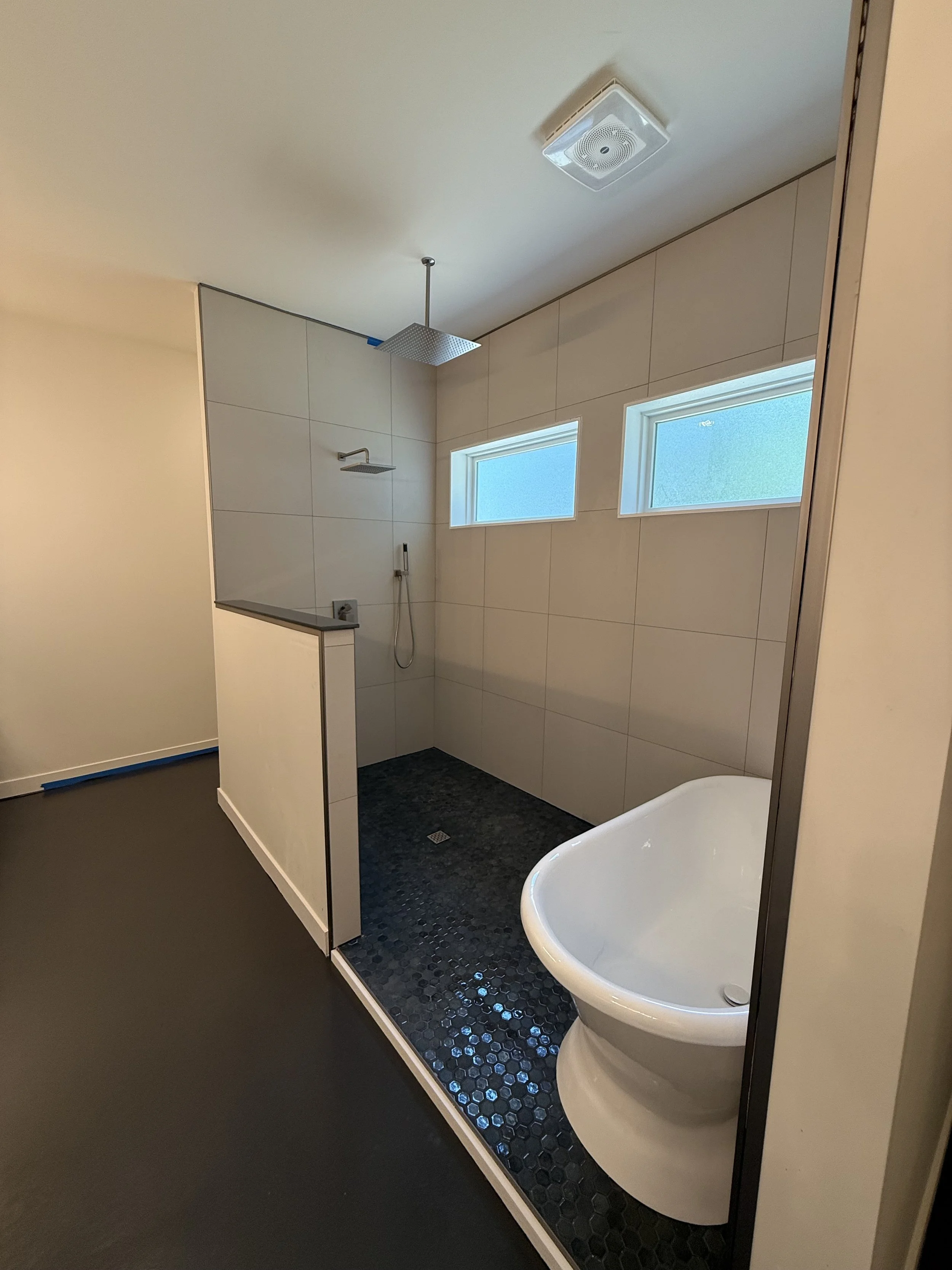 Modern bathroom with a walk-in shower area featuring two horizontal windows, black hexagonal floor tiles, a white corner bathtub, overhead shower head, and a ceiling vent.