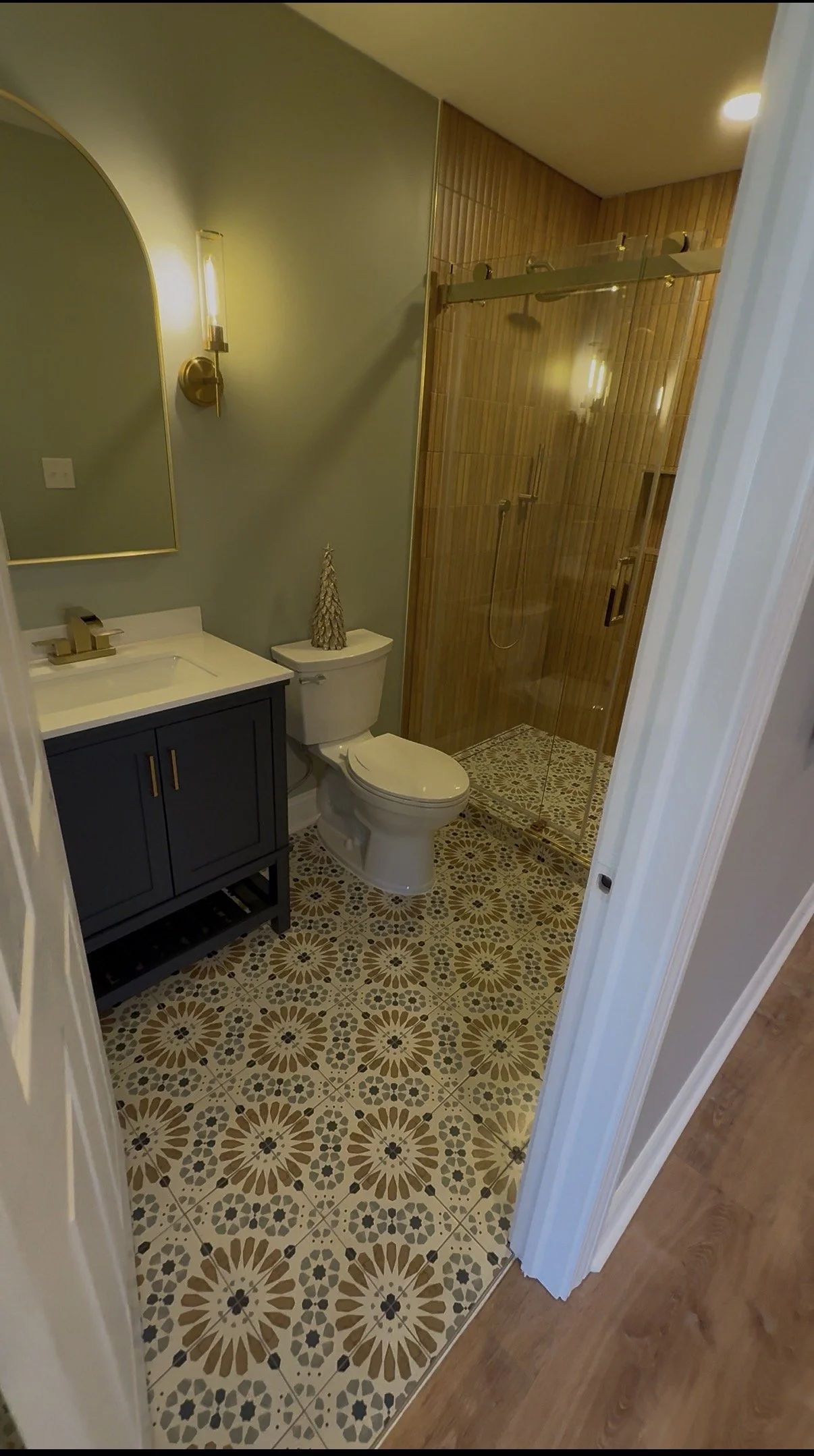 A small bathroom with a patterned tile floor, a navy vanity with a white countertop, a toilet, a gold mirror, a wall sconce light, and a glass-enclosed shower with wooden wall tiles.