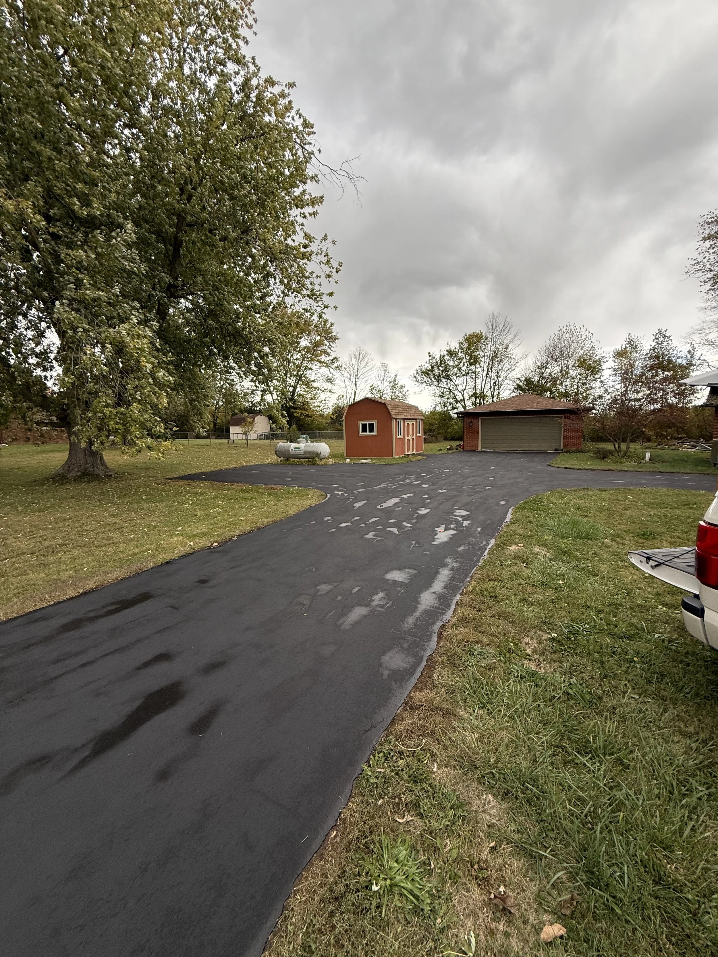 A freshly paved black asphalt driveway leading to a garage and shed in a residential yard. The yard has green grass, trees, and a cloudy sky overhead.