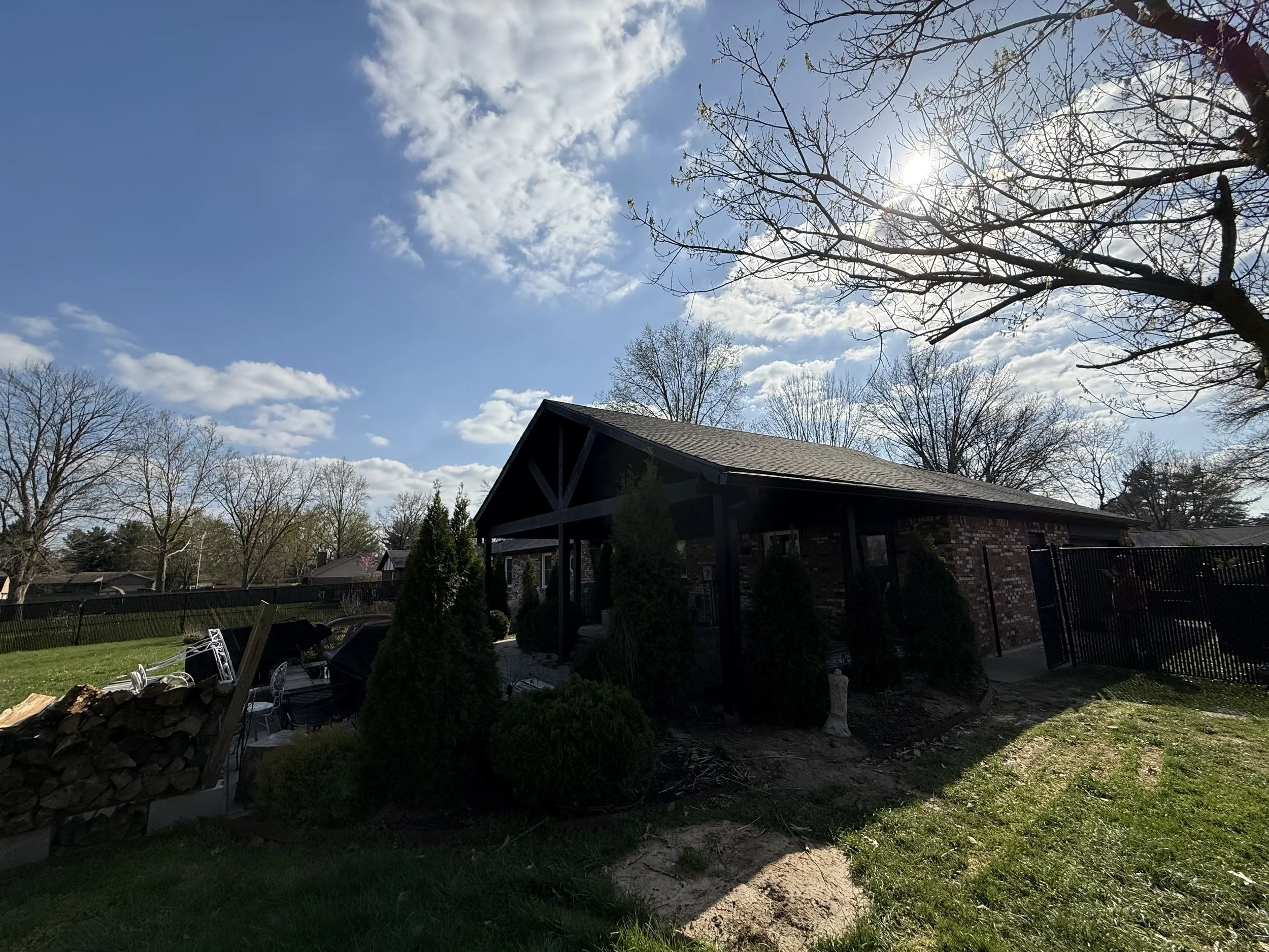 Backyard with a brick house, black fence, trees without leaves, a lawn, and a pile of chopped wood on the left side. The sky is partly cloudy and the sun is shining.