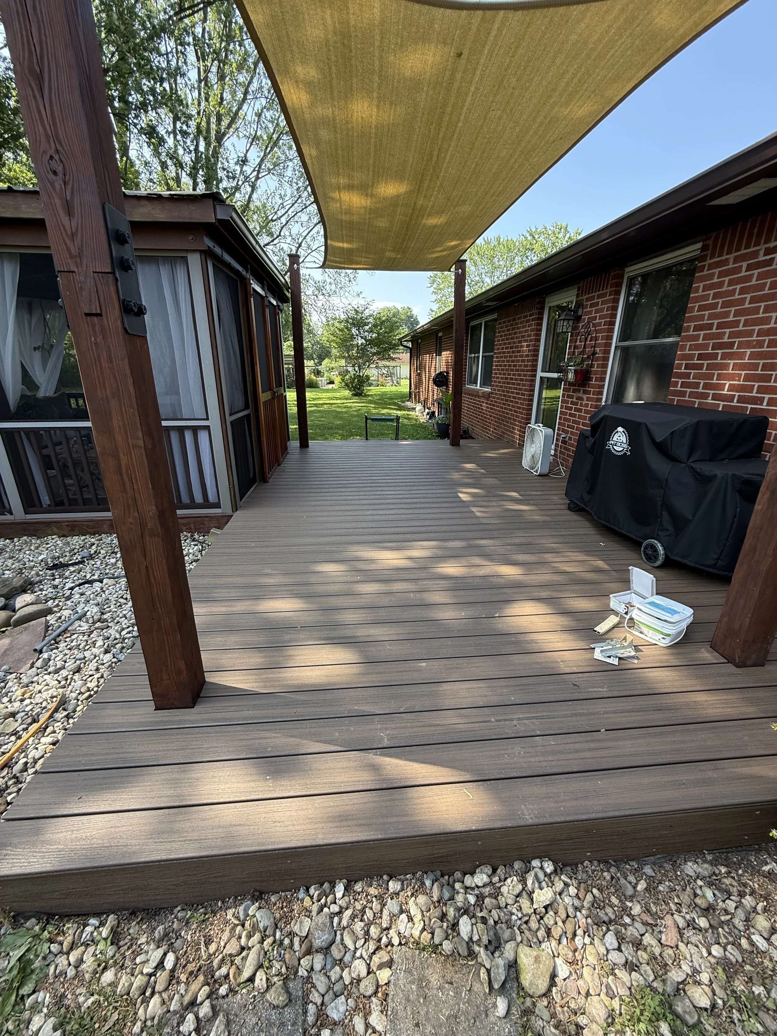 A wooden deck attached to a brick house with a railing, shaded by a yellow canopy, with a yard, trees, and a blue sky in the background.