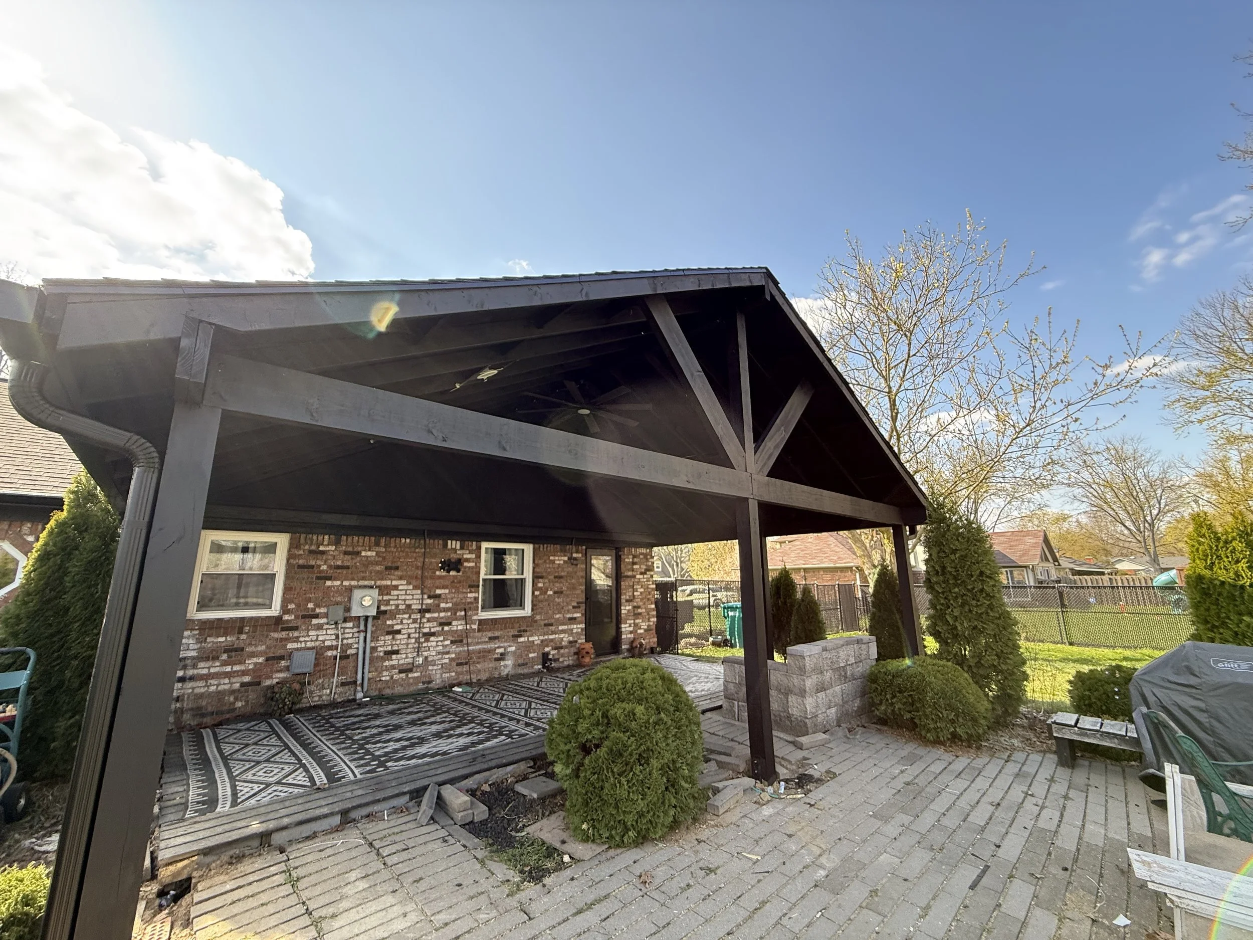 Backyard patio area with a black wooden pergola, brick house, outdoor rug, bushes, trees, and a clear blue sky.