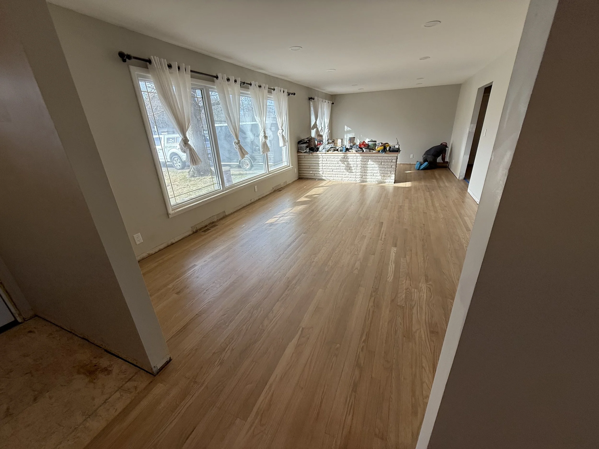 Empty living room with large windows, white curtains, and a worker installing a brick half-wall, with some tools and supplies on top.