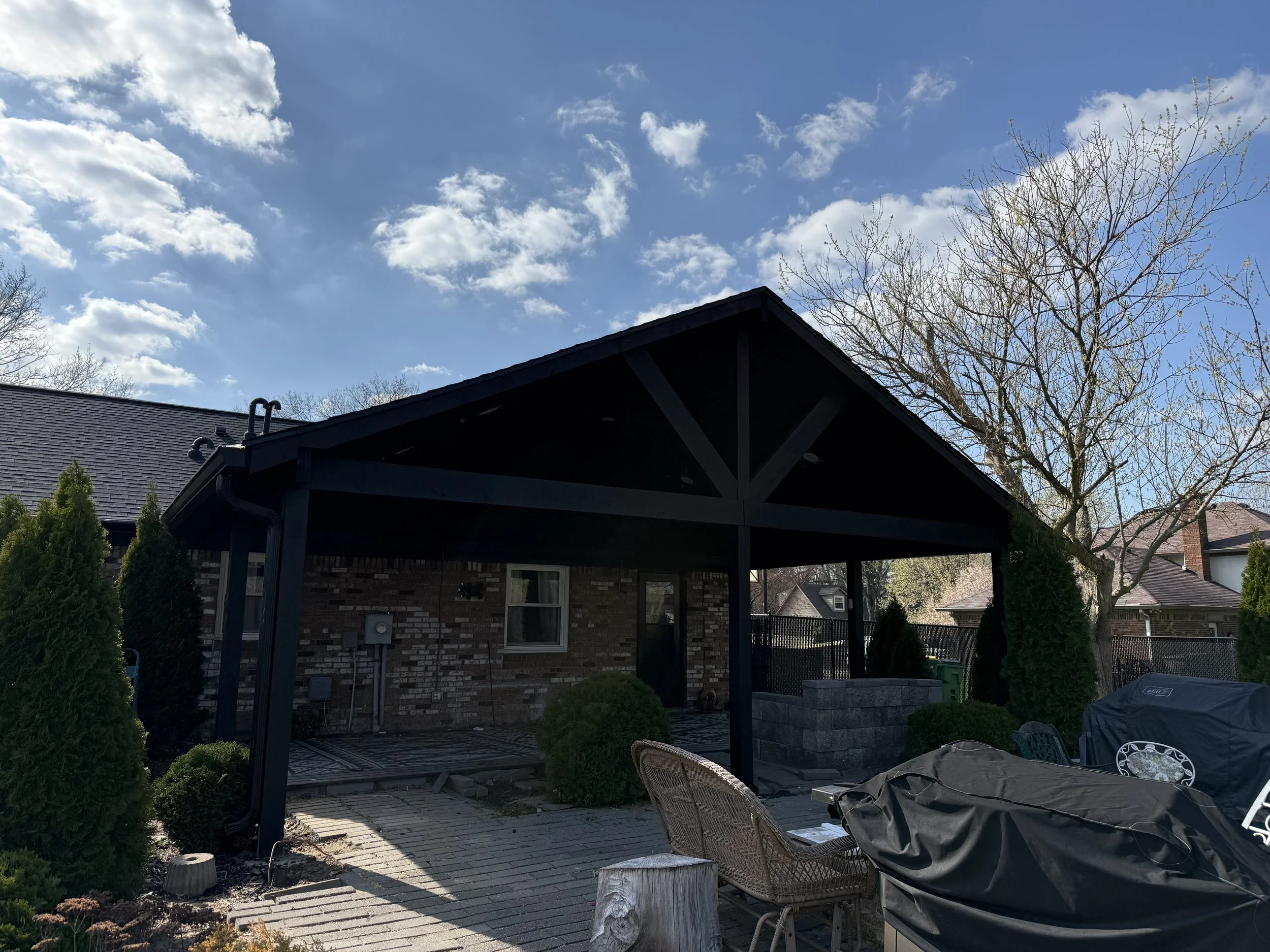 Backyard patio with a black covered porch, surrounded by bushes and trees, with outdoor furniture, a grill, and a brick house in the background under a partly cloudy sky.