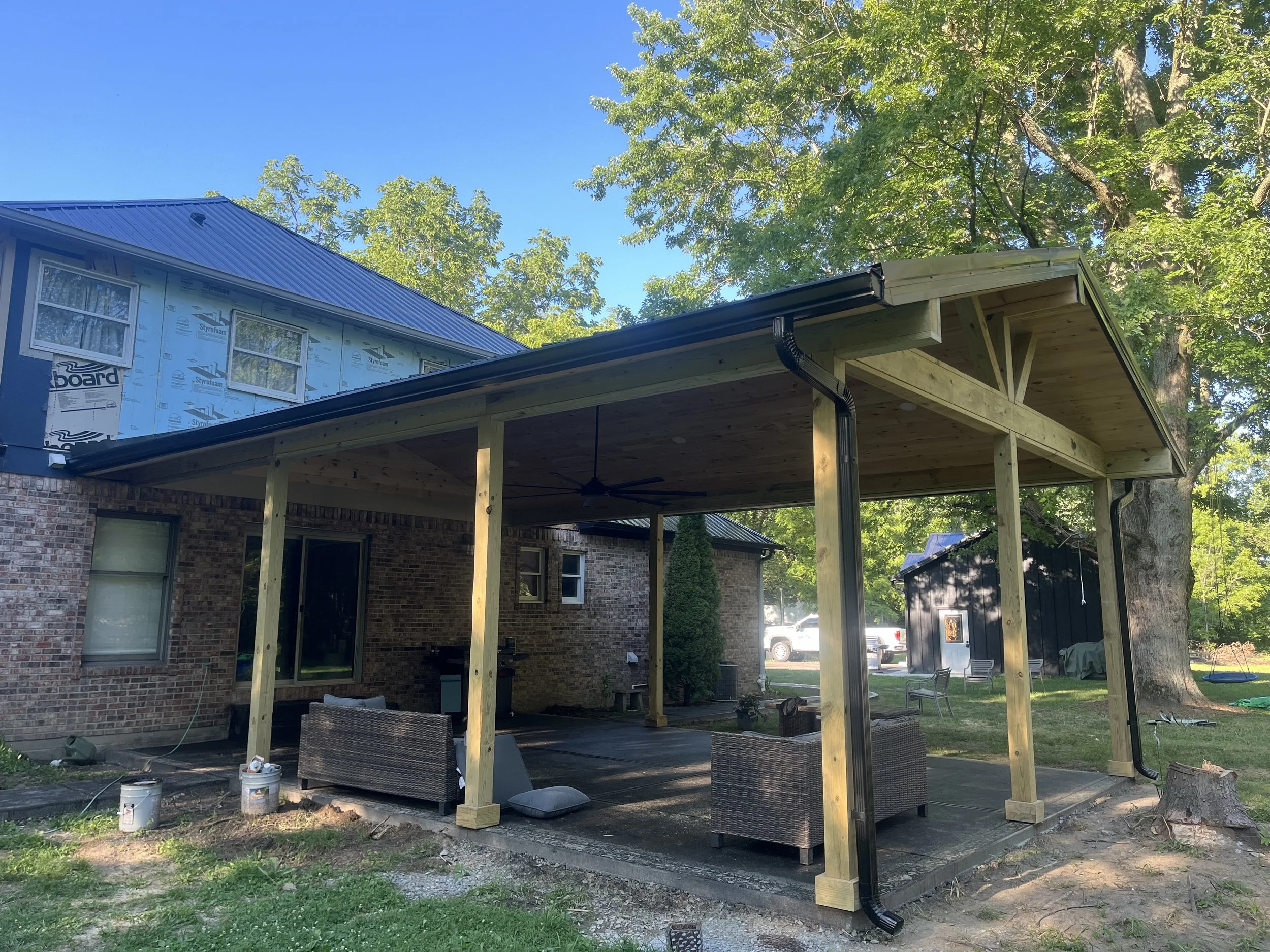 A brick house with a new wooden porch extension under construction, with construction materials and outdoor furniture, surrounded by green trees and a clear blue sky.