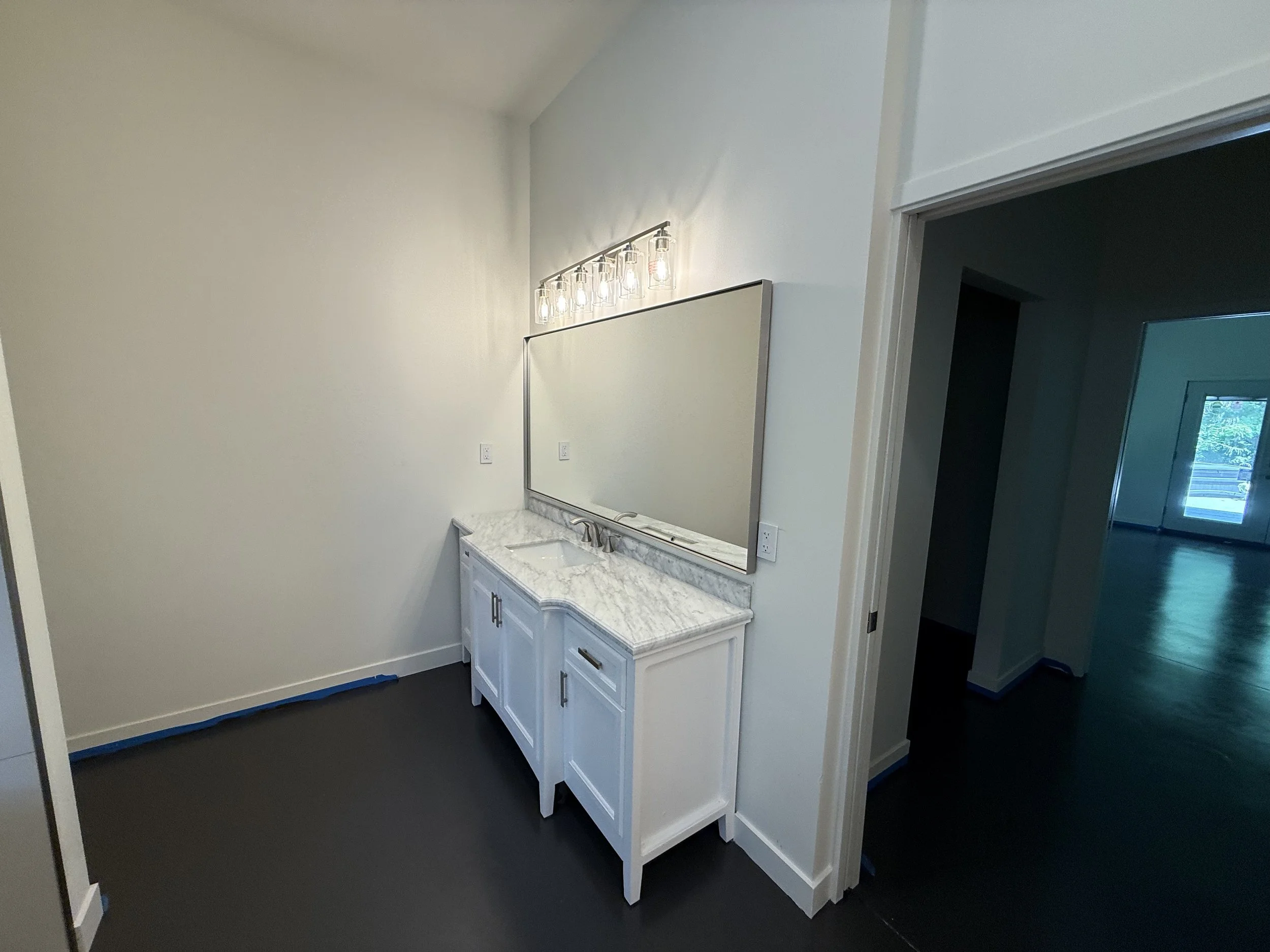 Bathroom vanity with a large mirror, white cabinets, and a marble countertop with a single sink, illuminated by a vanity light fixture, with an open doorway leading to a room with wood flooring and a window.