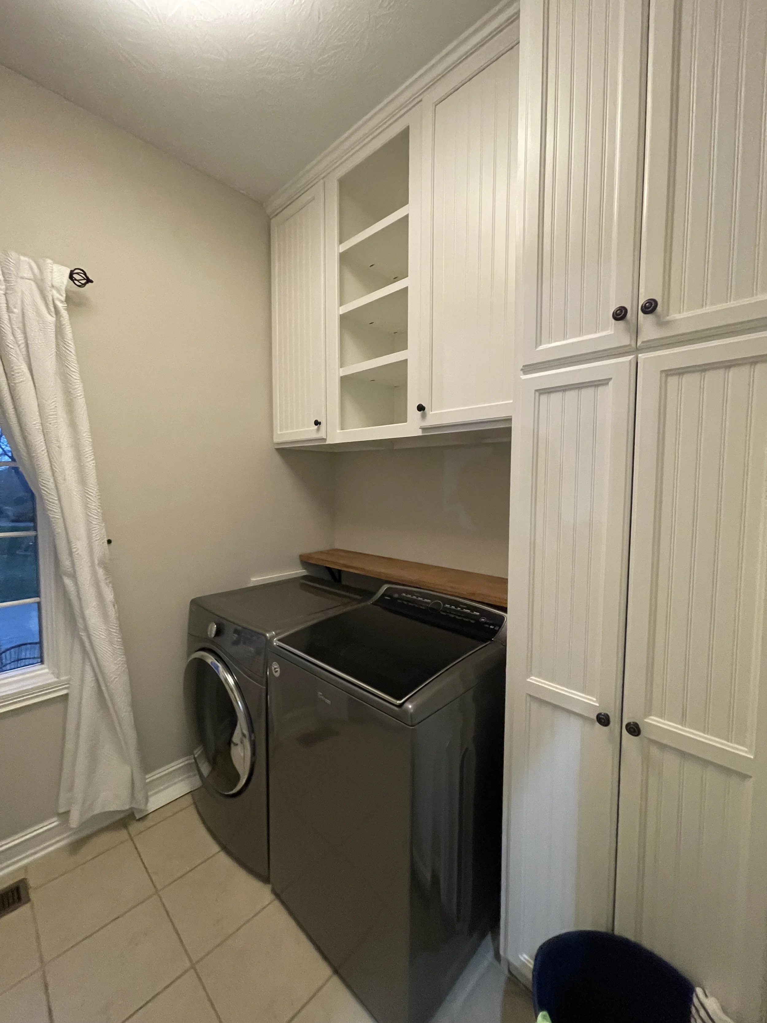 Laundry room with a front-loading washing machine and a top-loading dryer, white cabinets above, a window with curtains, and a small wooden countertop.