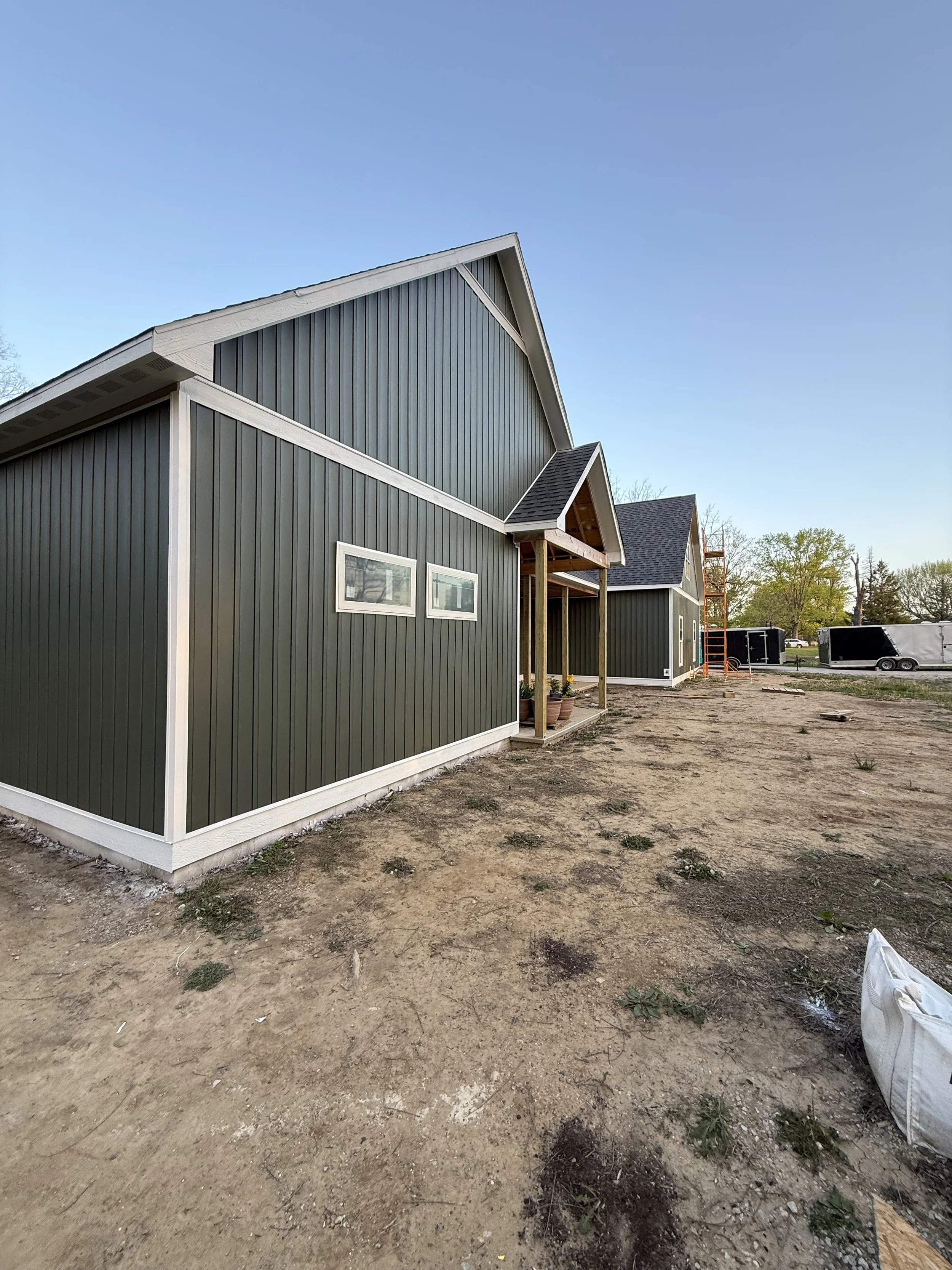 New house under construction with dark green siding, white trim, and small rectangular windows, set on a dirt lot with construction materials and trailers nearby.