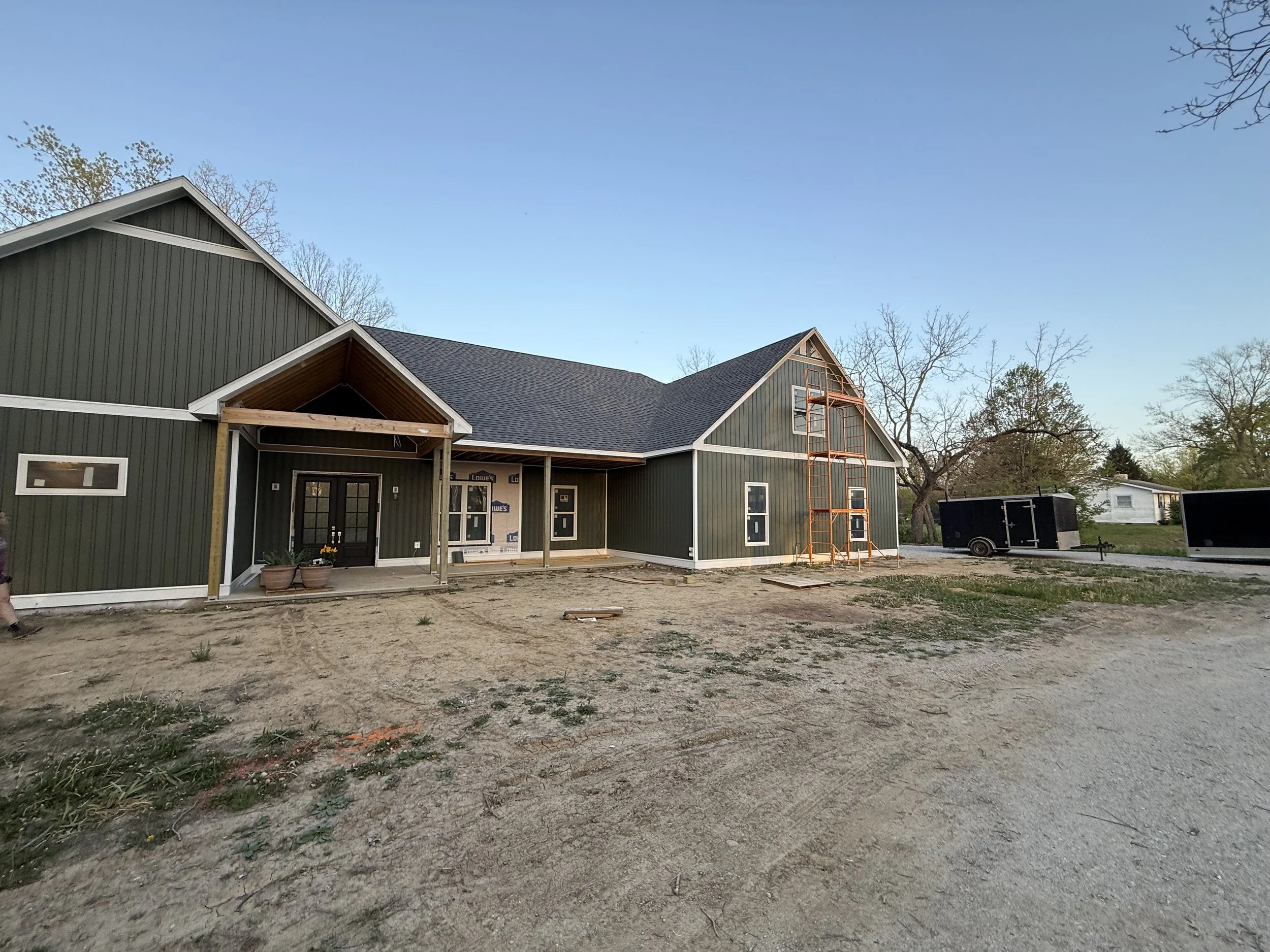 House under construction with green siding, a partially finished porch, and orange scaffolding on the right side. Construction materials and trailers are in the yard.