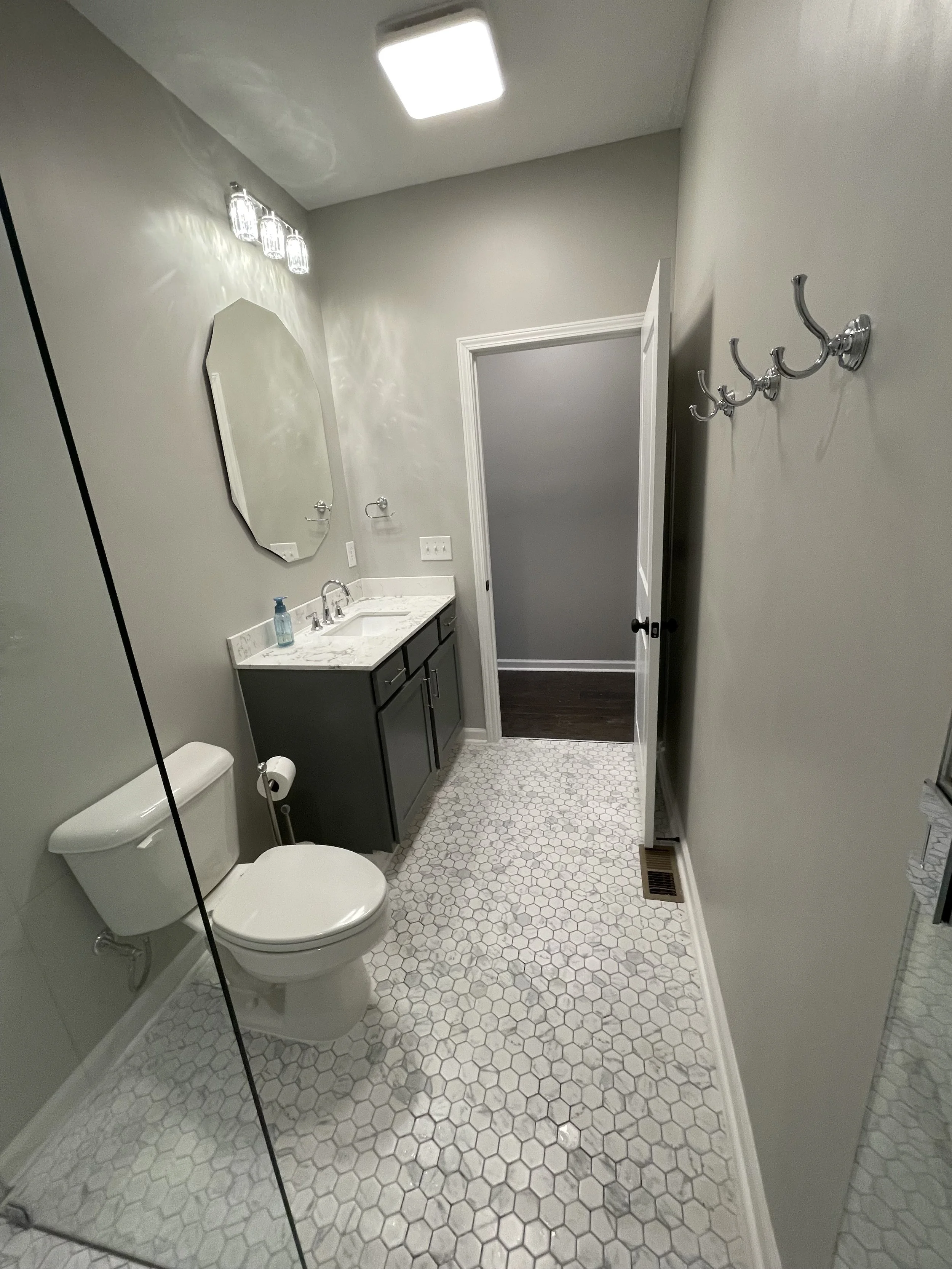 A small bathroom with a white toilet, dark gray vanity with a marble countertop, and a hexagon tile floor. A mirror above the sink, a light fixture, and a wall hook rack are also visible.