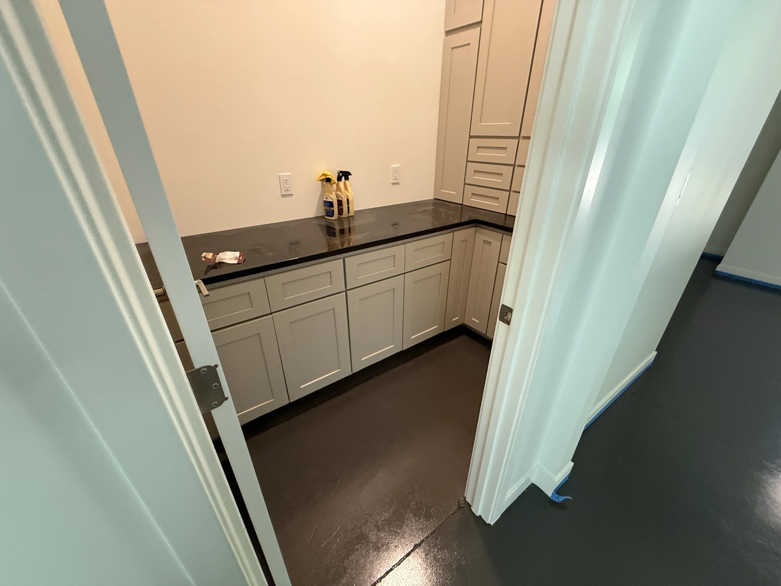 Empty kitchenette with beige cabinets, black countertop, and cleaning supplies on the counter.