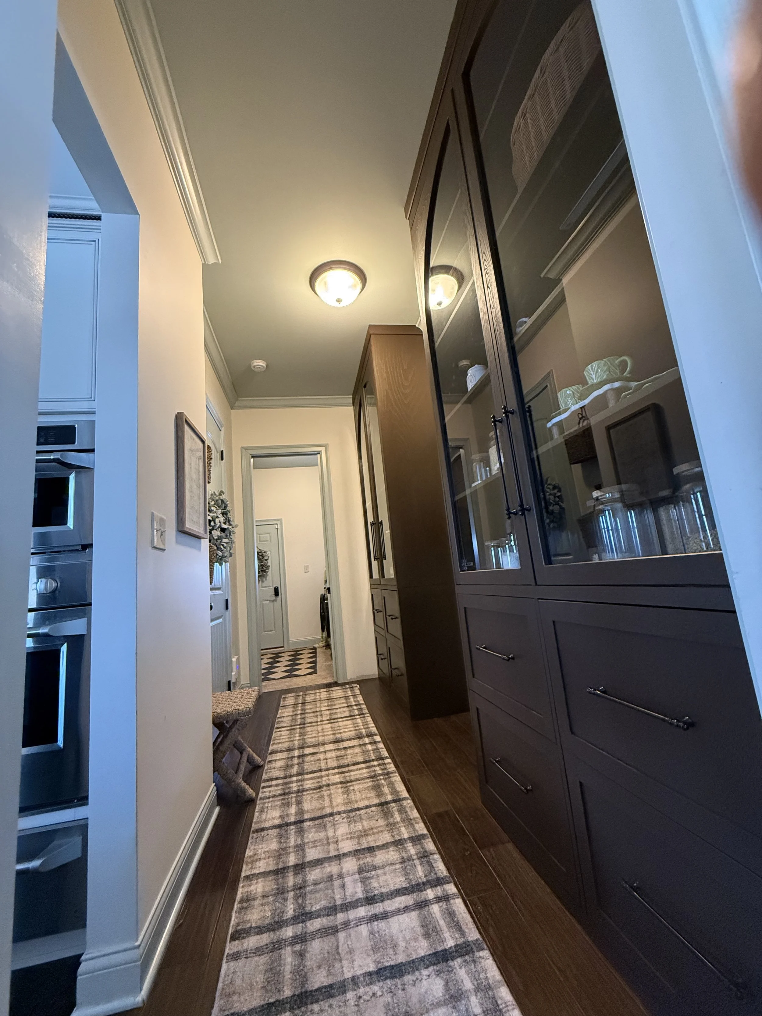 A narrow hallway in a house with dark wooden floors and beige walls. There is a long runner rug with a checkered pattern. On the right side, a large dark wooden cabinet with glass doors contains jars and dishes. At the end of the hallway, there is a 