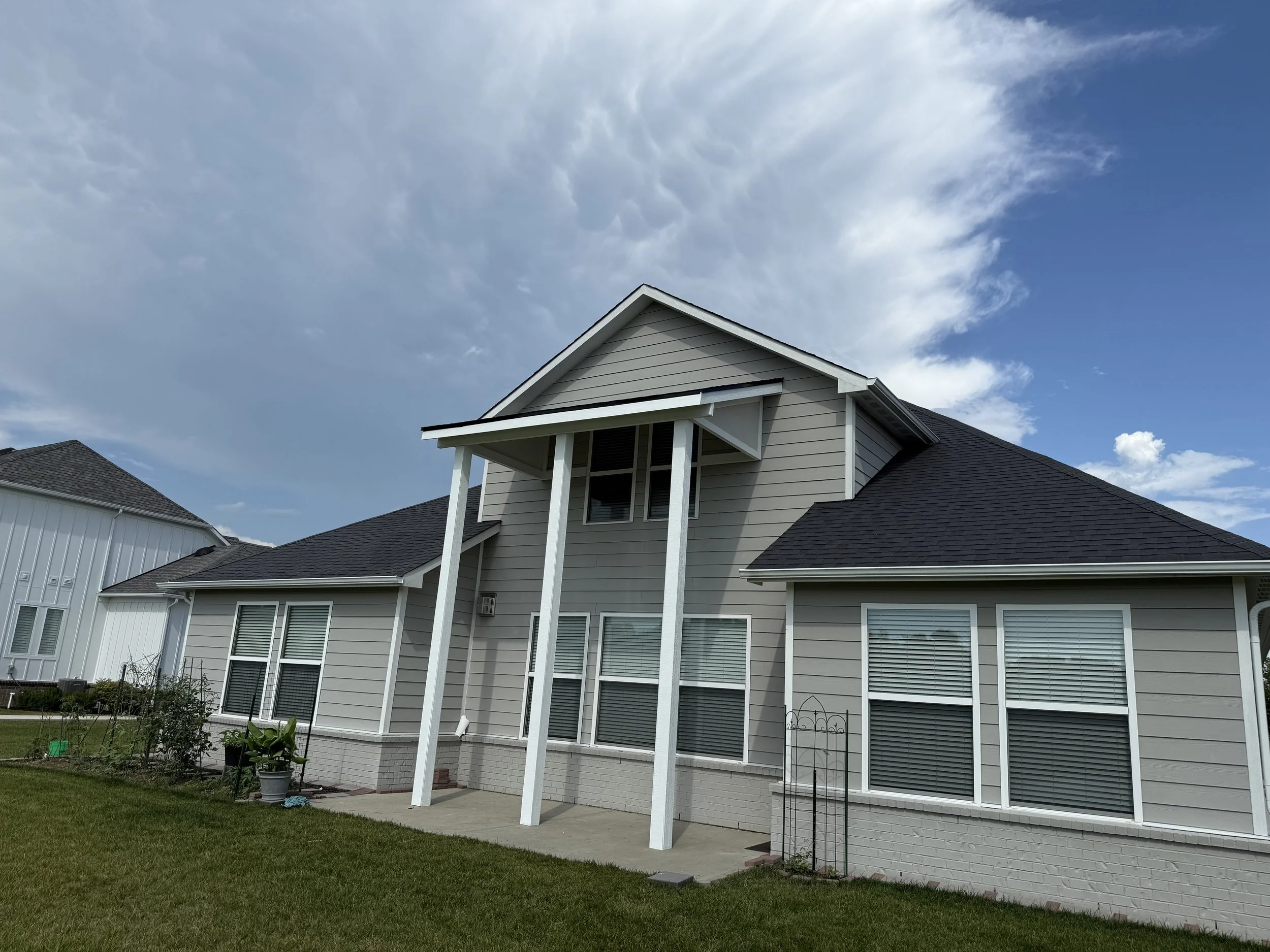 A two-story house with gray siding, white trim, and black roof shingles, featuring a small covered porch, multiple windows with blinds, a well-maintained lawn, and a partly cloudy sky in the background.