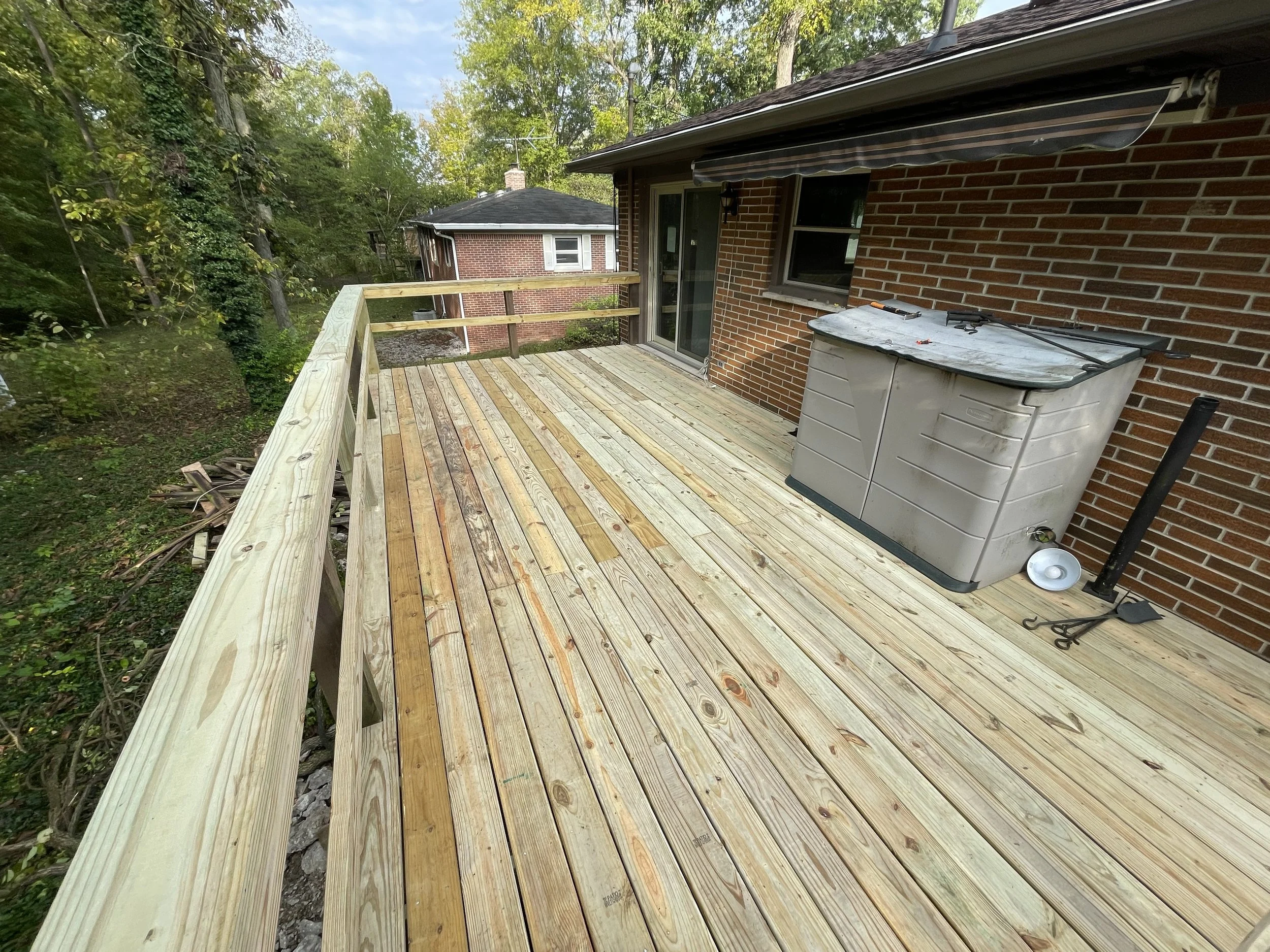 A newly constructed wooden deck attached to a brick house with a sliding glass door and window. The deck has light-colored wooden planks and a wooden railing. In the background, there are trees and another house. On the deck, there are some tools and