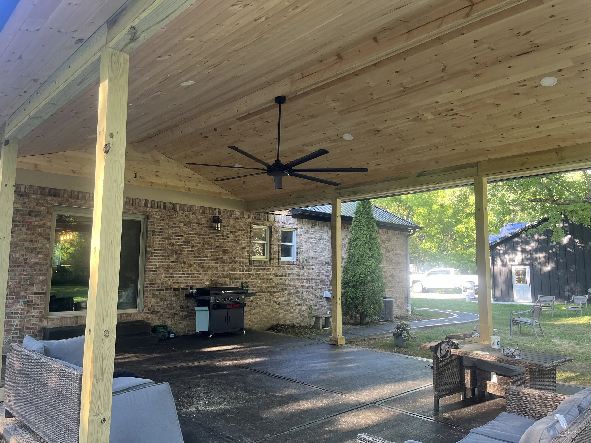 Covered outdoor patio area with a ceiling fan, brick wall, outdoor seating, and a grill, with trees and a small shed in the background.