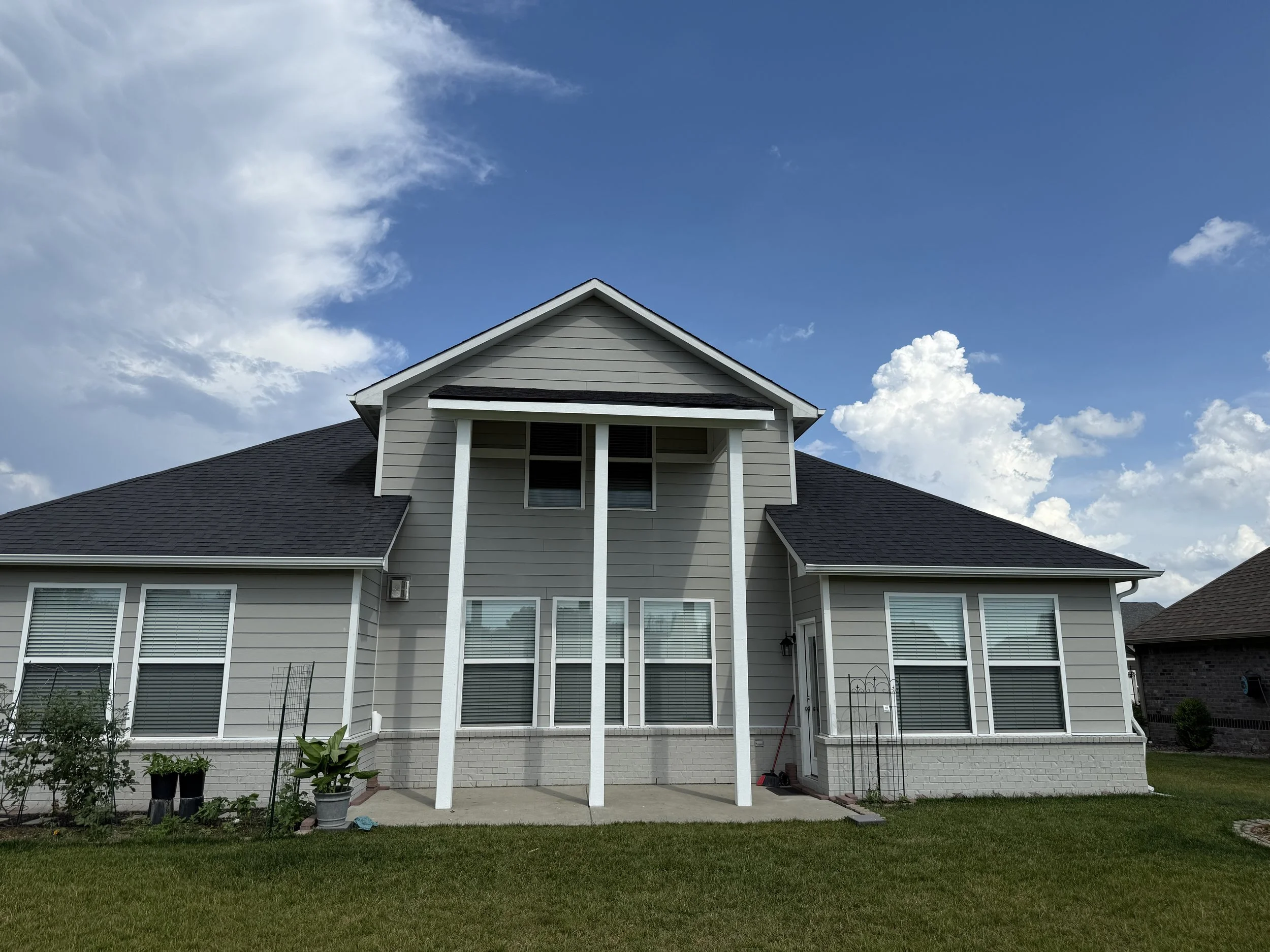 A two-story house with beige siding, white trim, and black roof, under a partly cloudy sky, with a small front yard and potted plants near the front porch.
