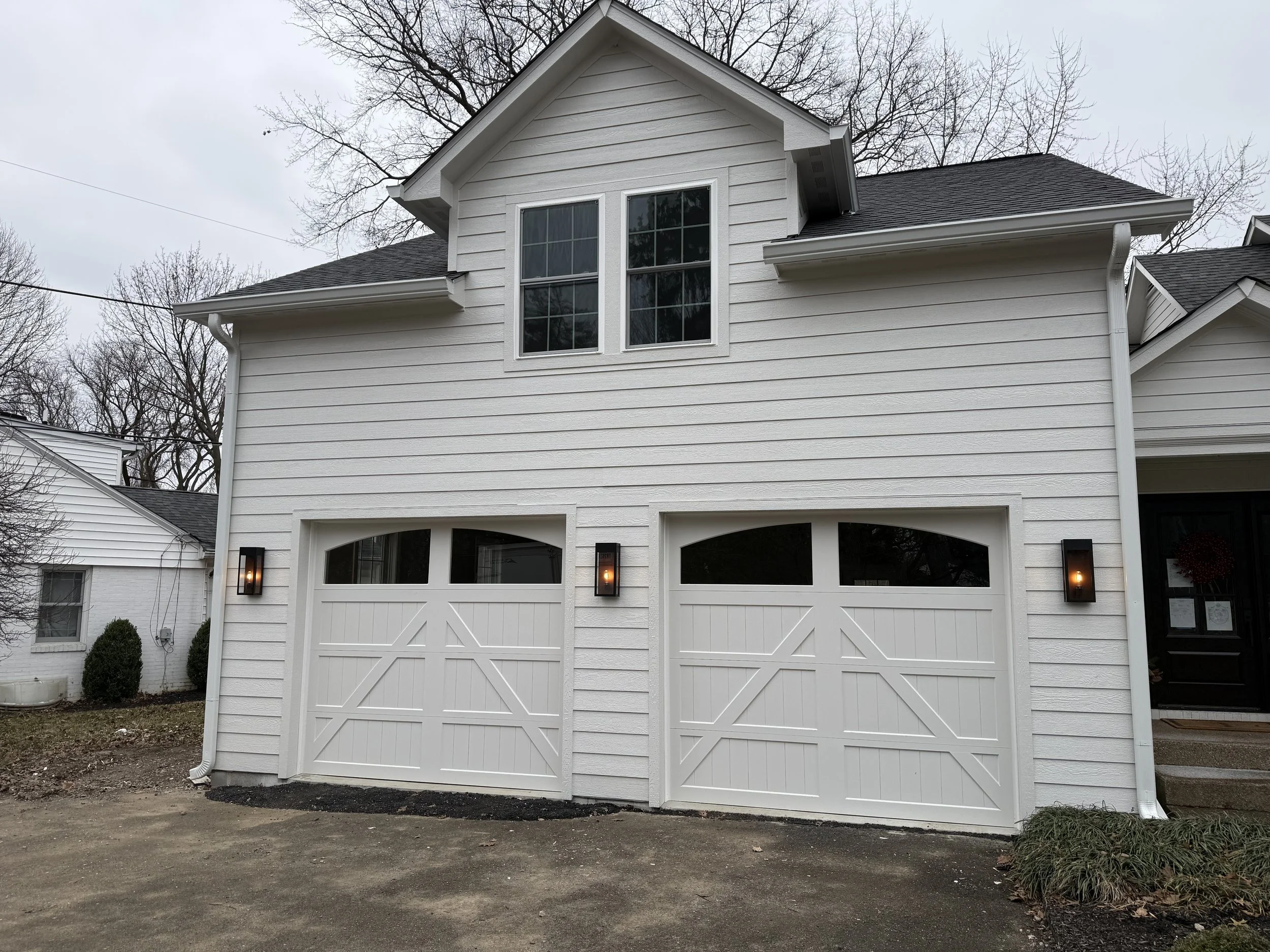 White two-story house with two-car garage, black lantern-style outdoor lights, and leafless trees in the background under cloudy sky.