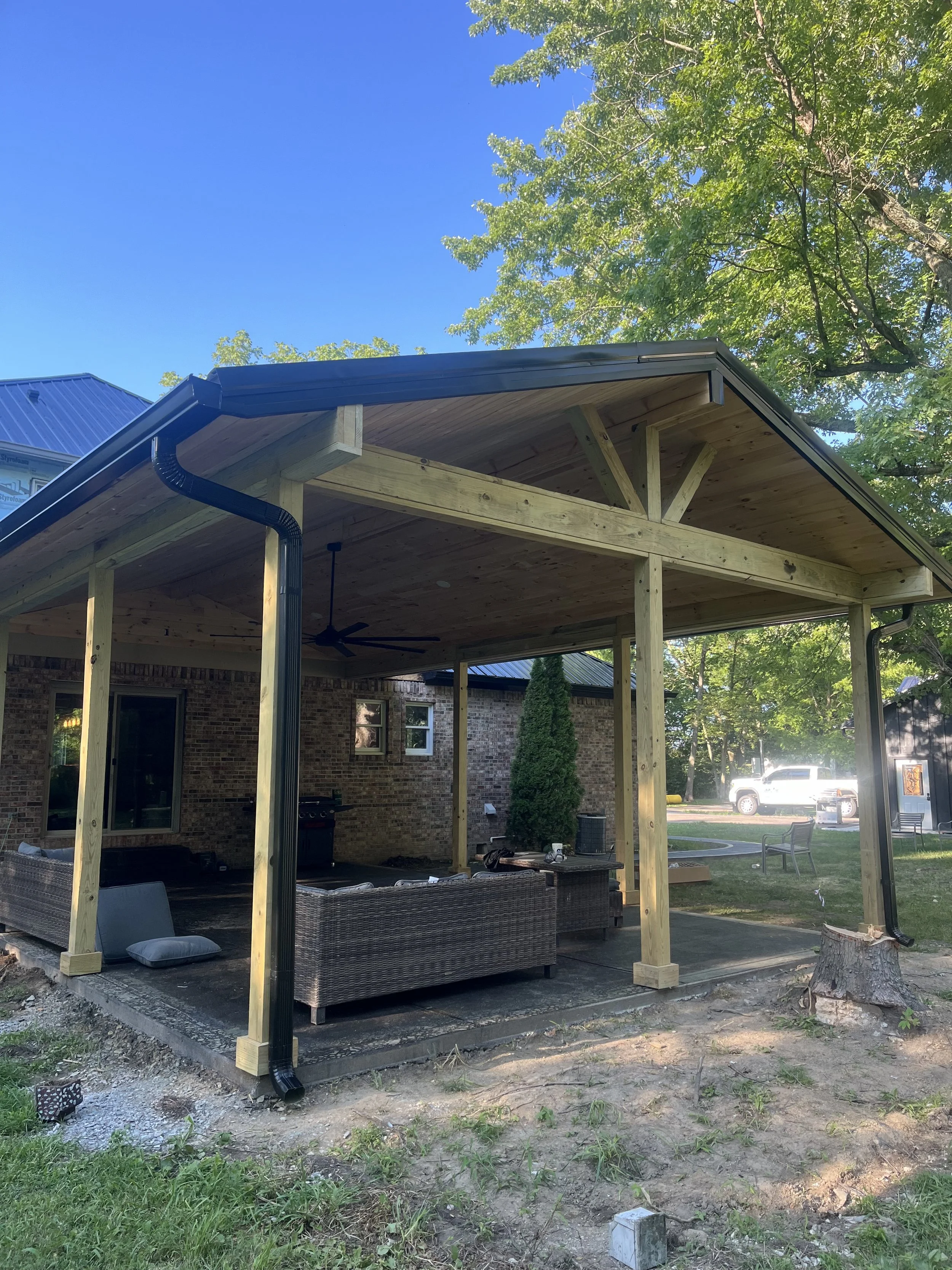 A backyard patio under construction with a wooden roof and wicker furniture, brick house in the background, and a truck parked in the distance surrounded by green trees.