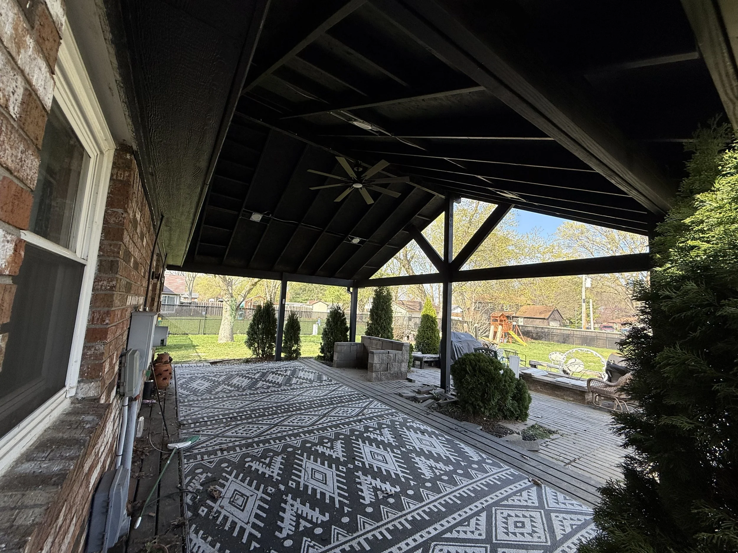 Covered backyard patio with patterned outdoor rug, some shrubs, a children's play set in the distance, and a grill covered in a tarp.