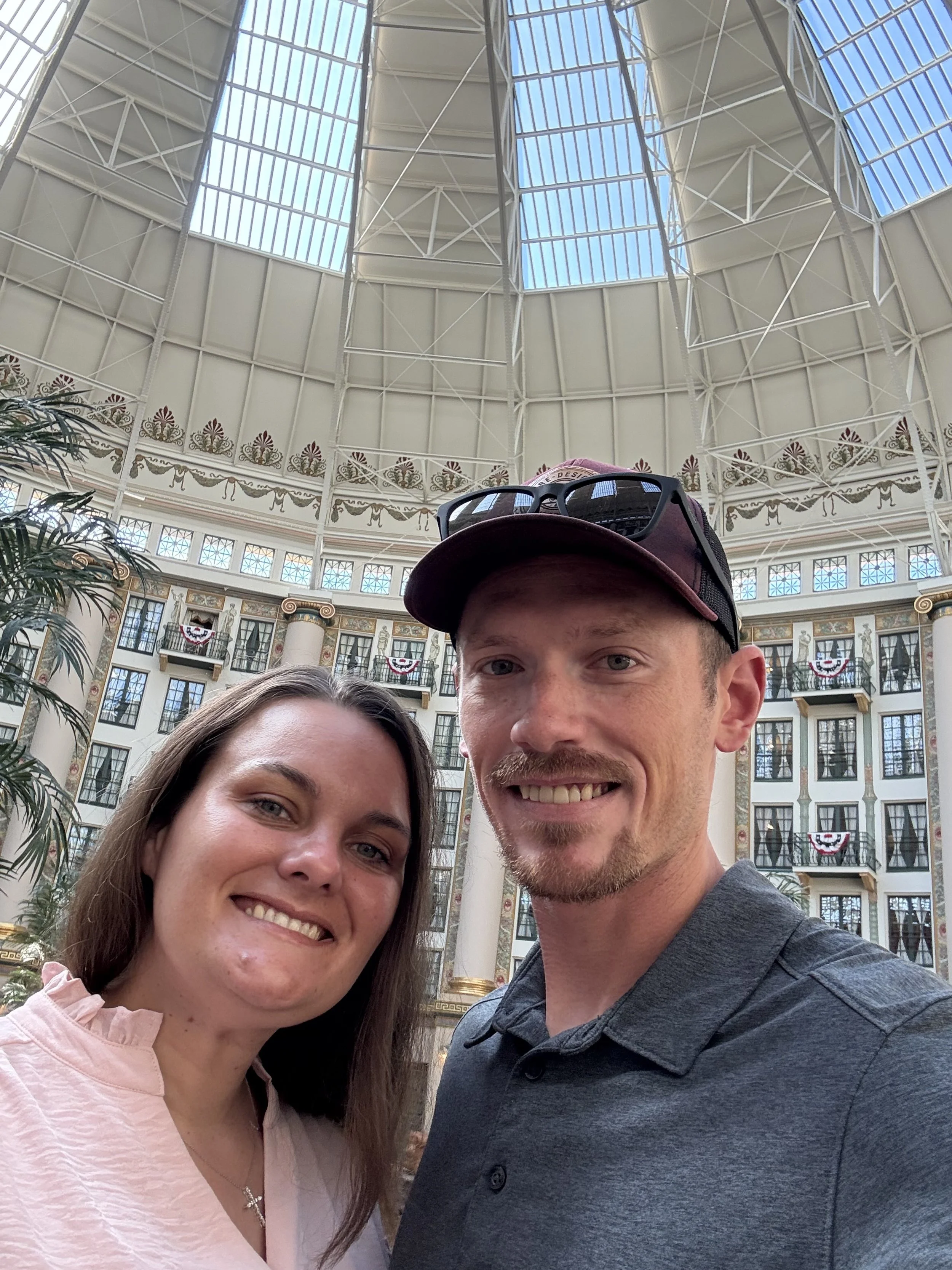 A smiling couple taking a selfie inside a grand, domed indoor atrium with ornate decorations, tall columns, and large windows at the top, with American flags hanging in the background.
