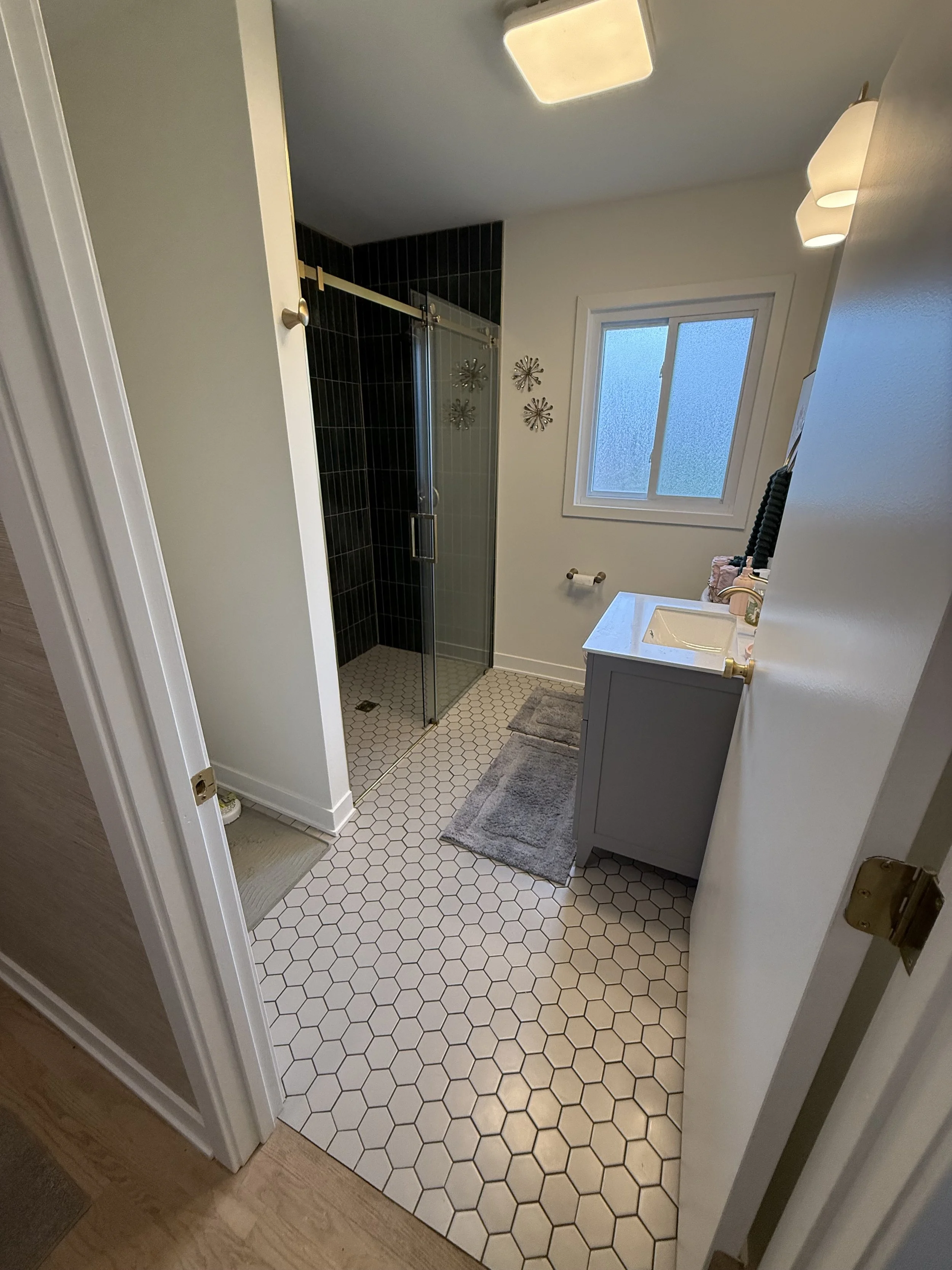 Bathroom with hexagonal tile floor, glass shower enclosure with black tiles, white vanity, and frosted window.