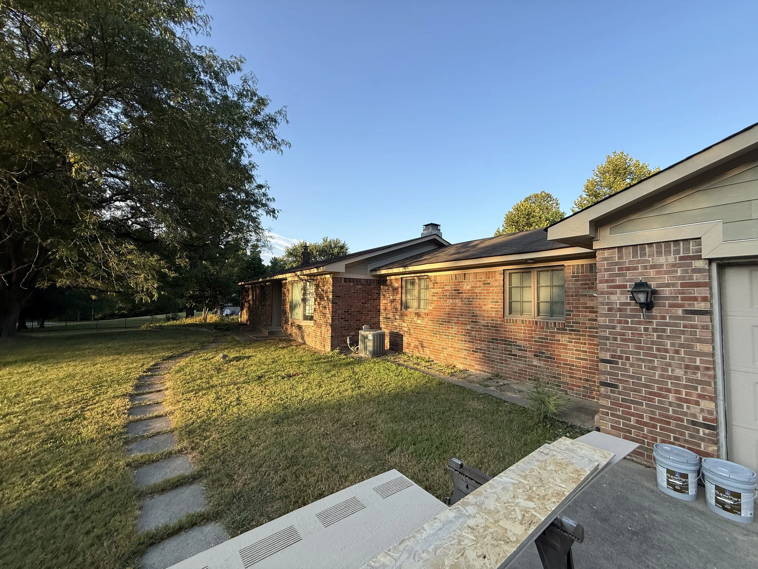 Side view of a brick house with a light-colored siding section, green lawn, stepping stones, a large tree on the left, and a clear blue sky overhead.
