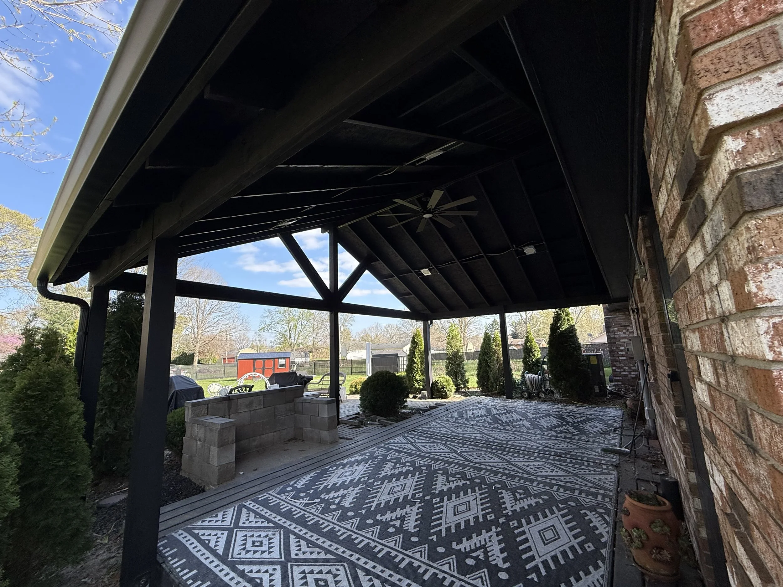 Covered outdoor patio with a patterned rug, brick wall, potted plants, and a yard with trees and a red shed visible in the background.