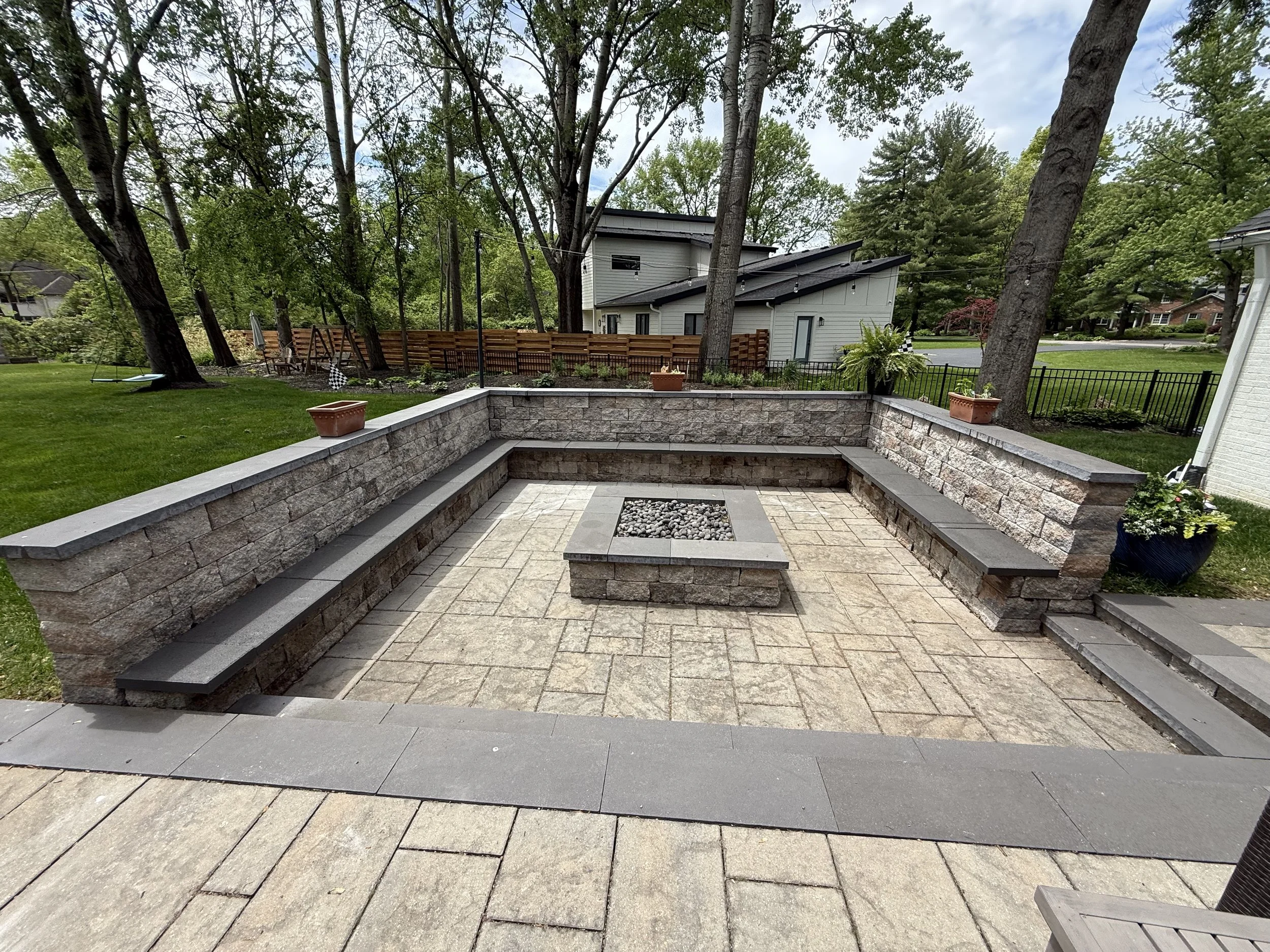 Backyard patio with stone retaining wall, built-in stone benches, and a small fire pit filled with pebbles, surrounded by trees and residential houses.