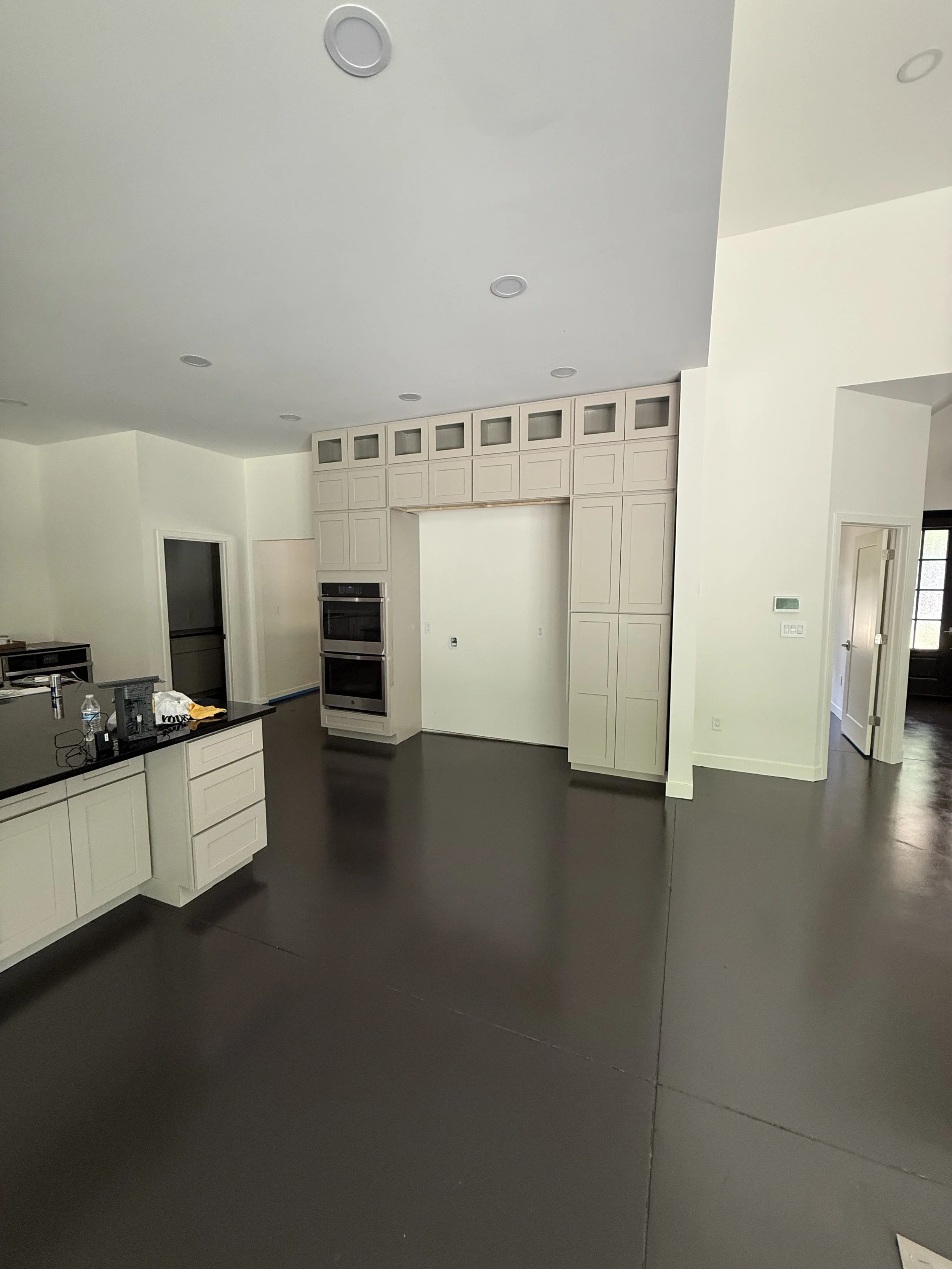 Empty kitchen with white cabinets, black countertop, and built-in double oven, dark hardwood flooring, and white walls.