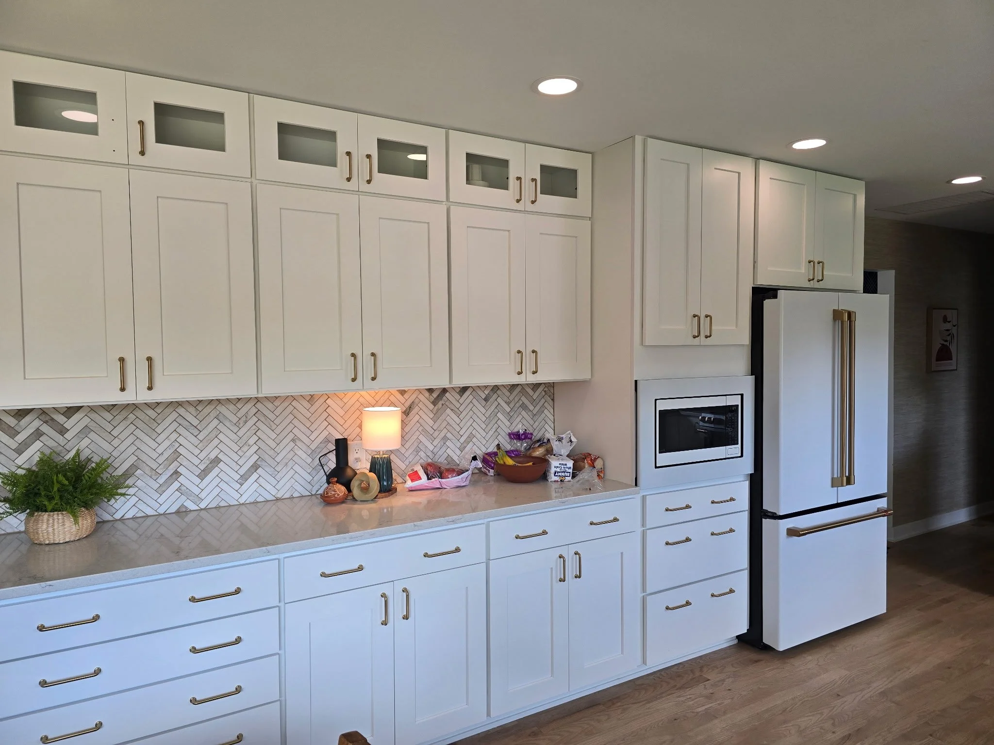 Modern kitchen with white cabinets, gold handles, built-in microwave, refrigerator with gold handles, backsplash with white herringbone tiles, small lamp, plants, and assorted decorative items on the countertop.