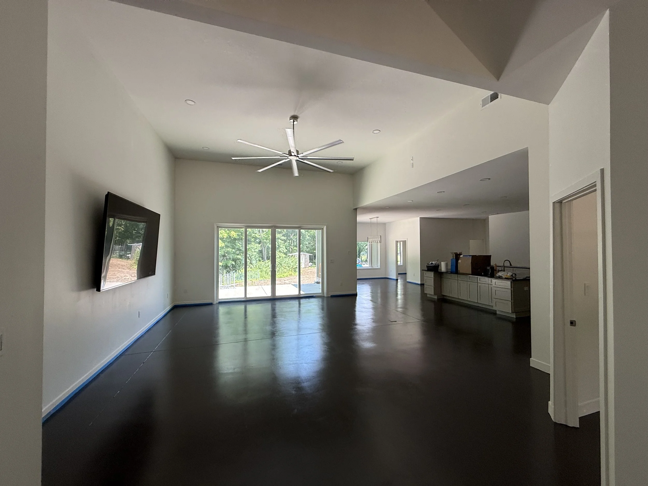 Empty living room with white walls, dark hardwood floors, a large sliding glass door, a ceiling fan, and a view of a green backyard; adjacent kitchen area with white cabinets.