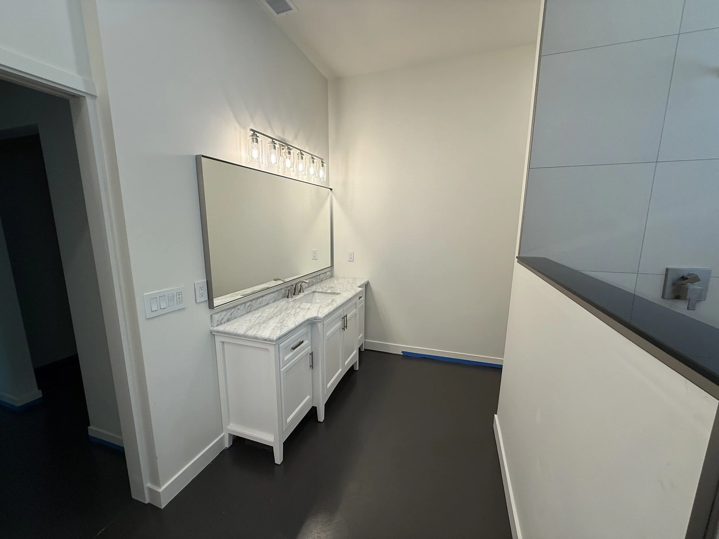 A small bathroom vanity with a white cabinet, a marble countertop, and a built-in sink, located beneath a large mirror and a light fixture with six exposed bulbs. The wall behind the vanity is painted white, and the floor is dark-colored.