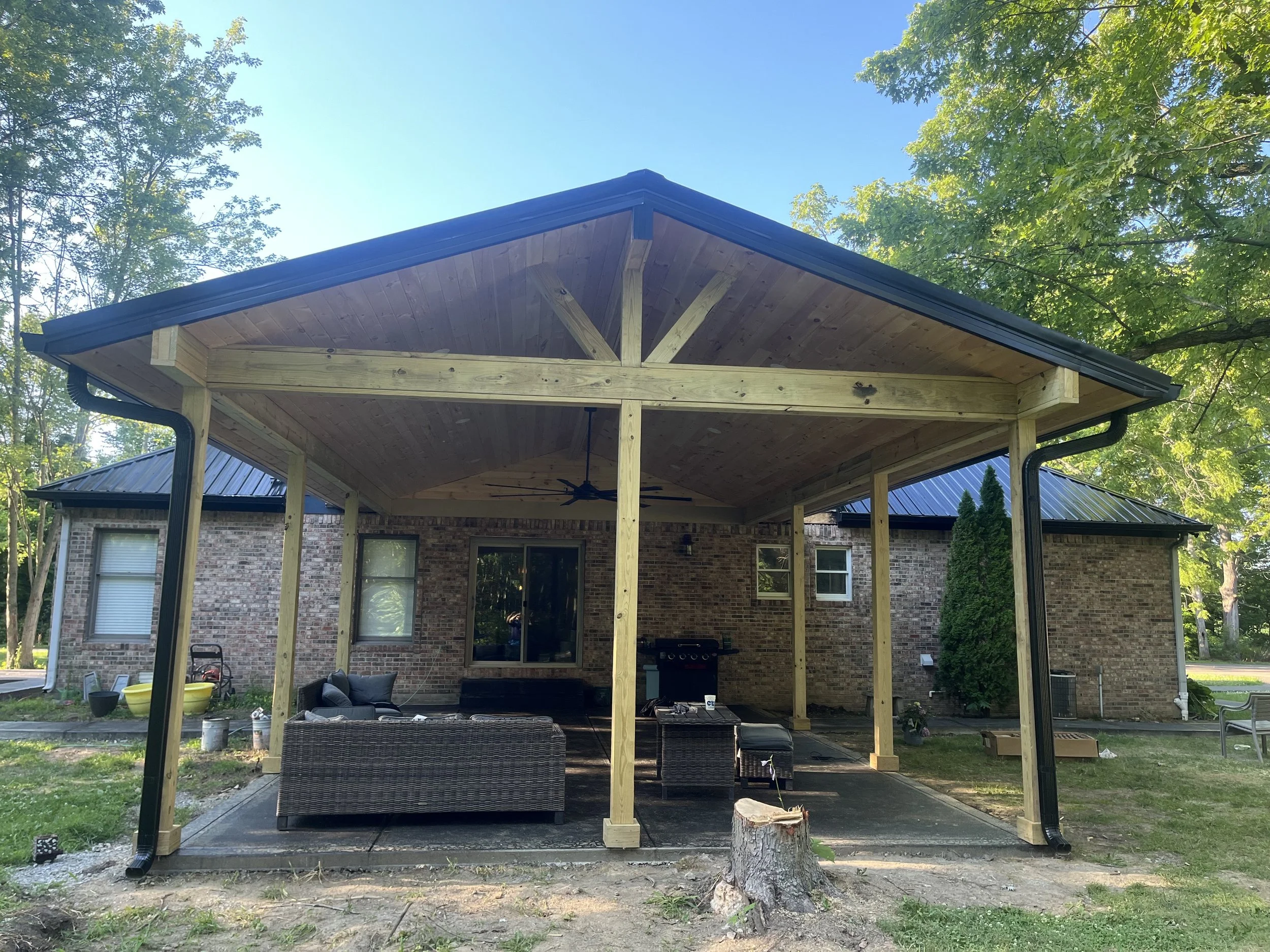 View of a backyard patio with a newly built wooden roof extension supported by wooden posts. The house in the background is made of brick, with visible windows and a sliding glass door. Outdoor furniture includes a couch and other seating, with trees