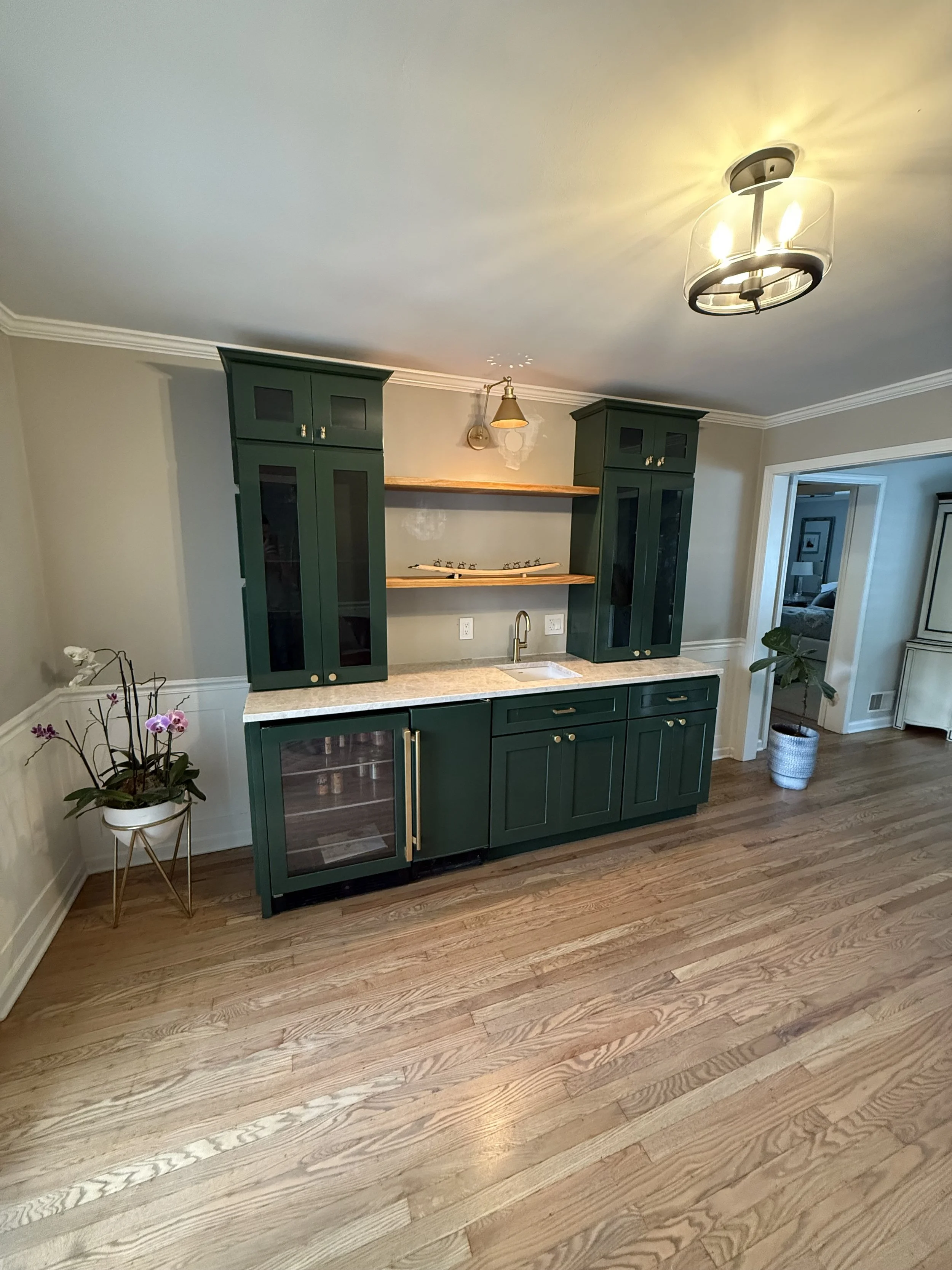 Green kitchenette with cabinets, open shelving, a small sink, and a mini fridge in a room with wooden flooring and ceiling lights.