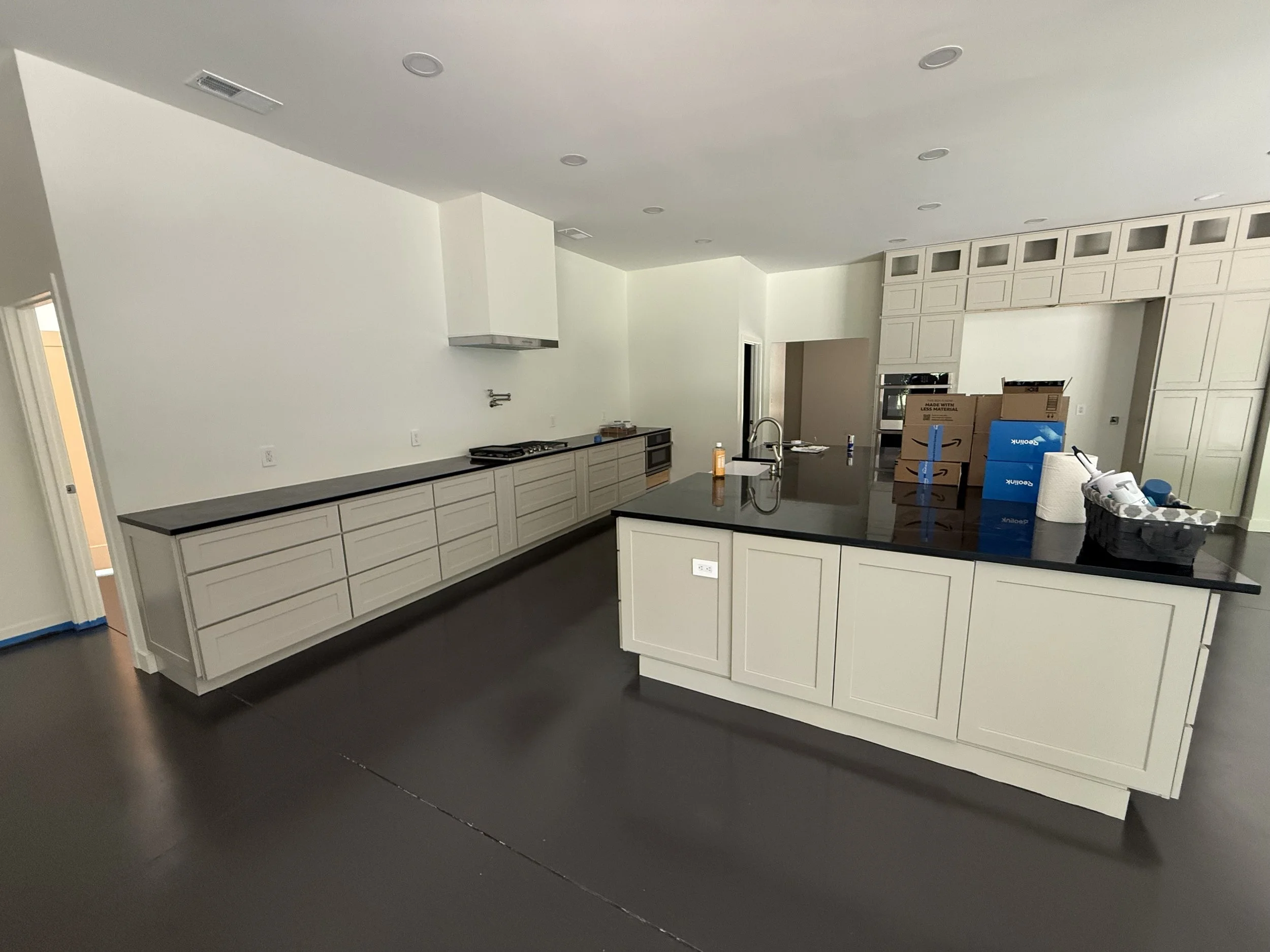 Empty kitchen with white cabinets, black countertops, and a large island with a sink. There are boxes and packing supplies on the island, suggesting ongoing unpacking or moving.