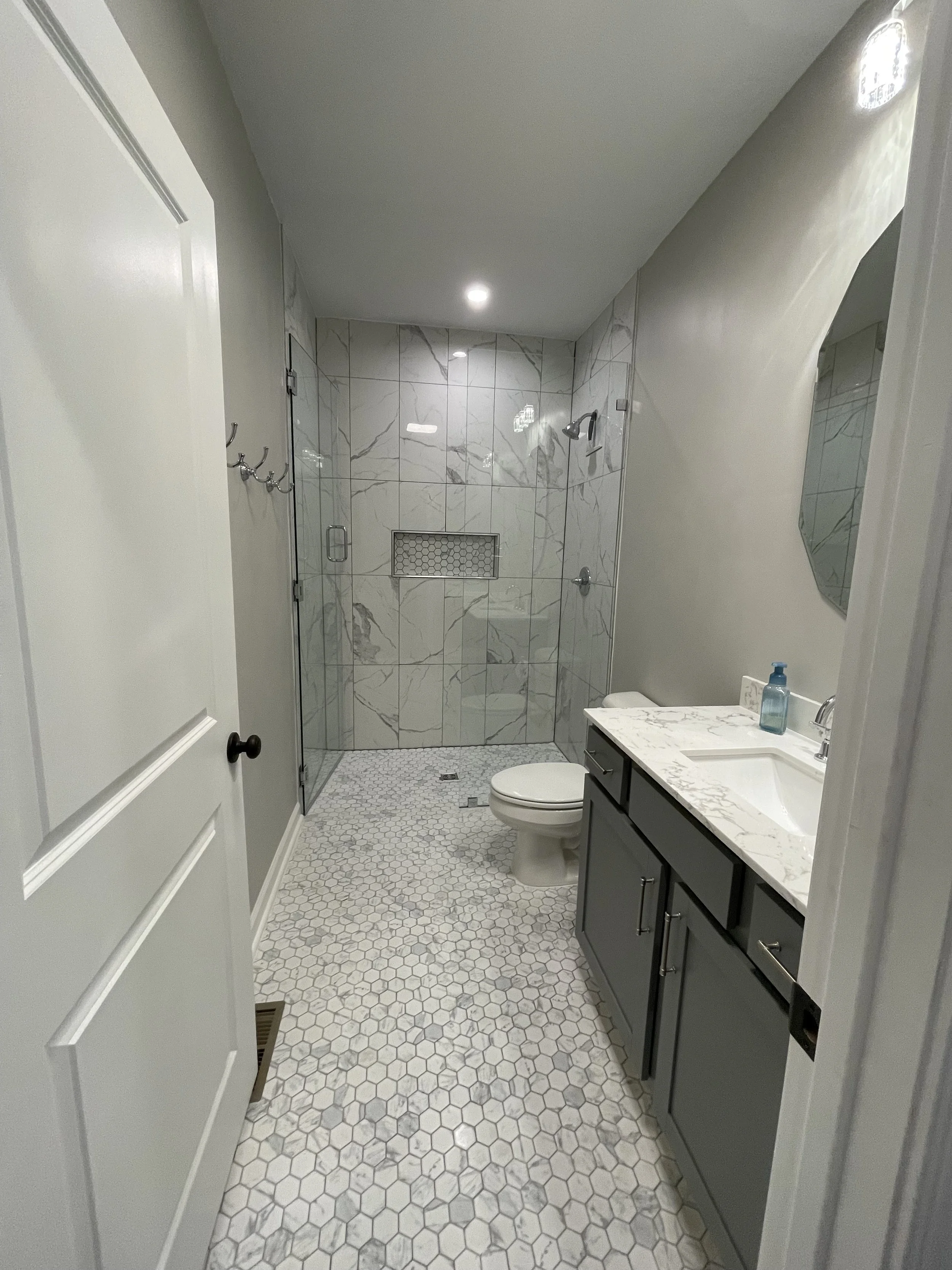 Bathroom with a walk-in shower, toilet, and vanity with a marble countertop, gray cabinets, and a hand soap dispenser.