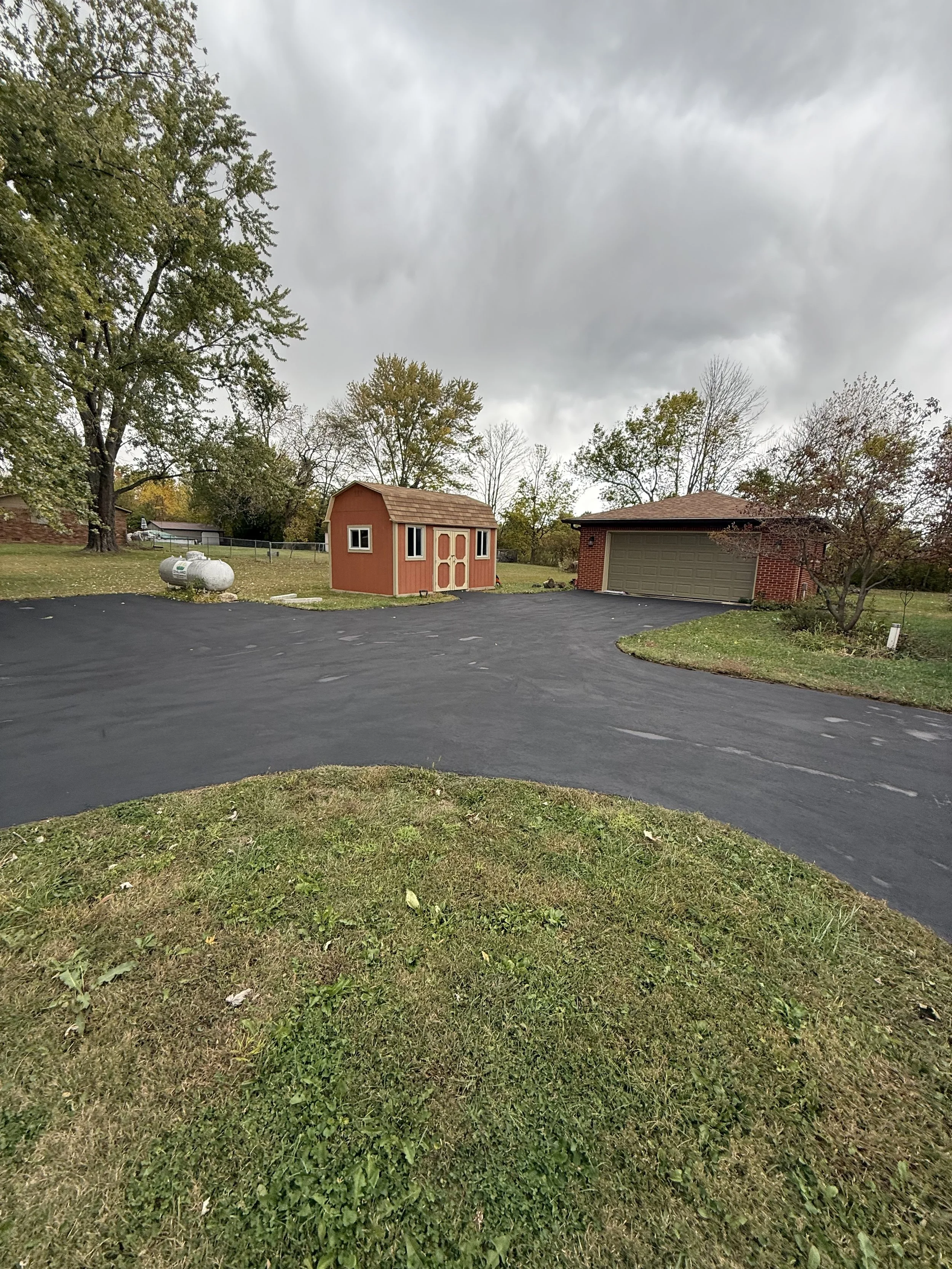 A residential driveway with a newly paved black asphalt surface, a small red shed with beige trim, a brick garage with a green door, and trees with some autumn foliage under a cloudy sky.