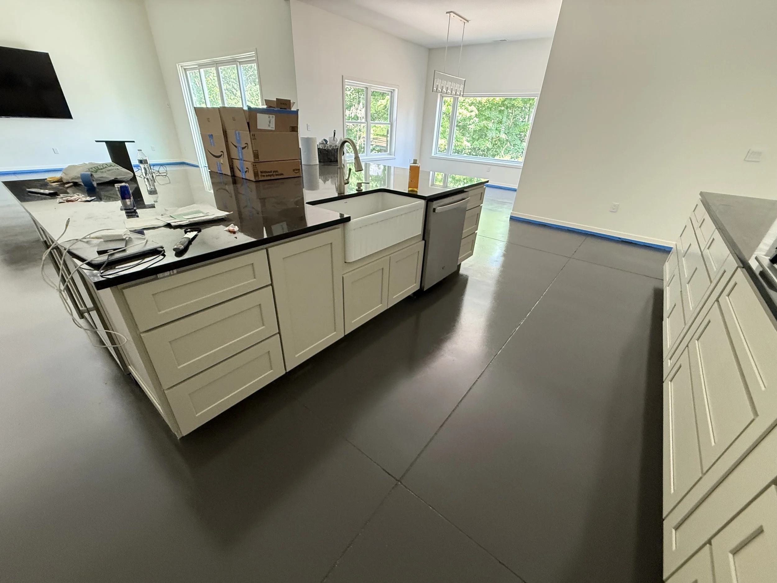 Kitchen with white cabinets, black countertops, a large island with a farmhouse sink, and stainless steel dishwasher. The floor is unfinished concrete, and blue painter's tape lines the edges of the walls. There are construction materials and tools o