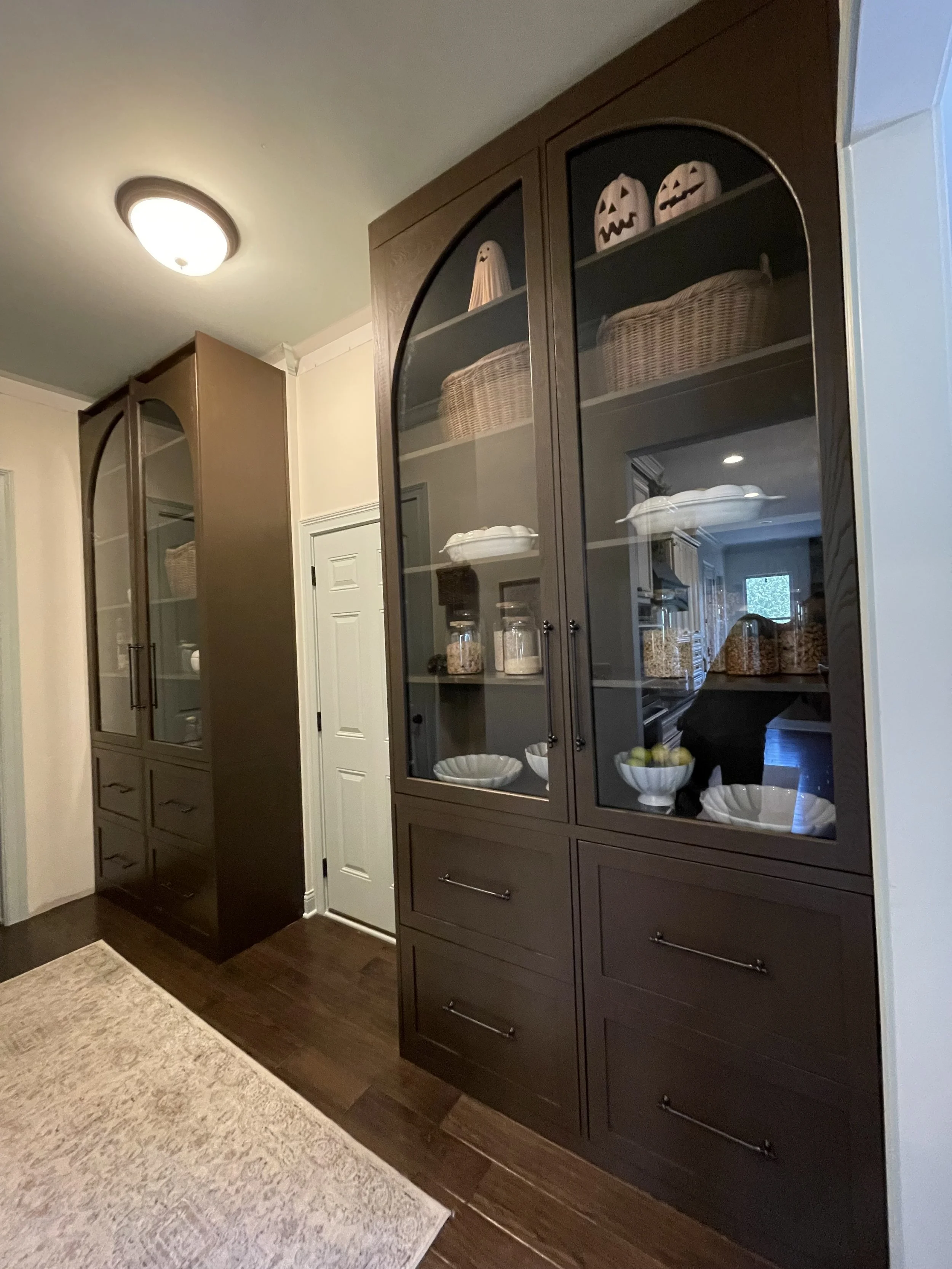 View of a kitchen corner with two dark wood cabinets with glass doors displaying Halloween decorations, jars, and bowls, next to a white door and a beige rug on dark hardwood floor.