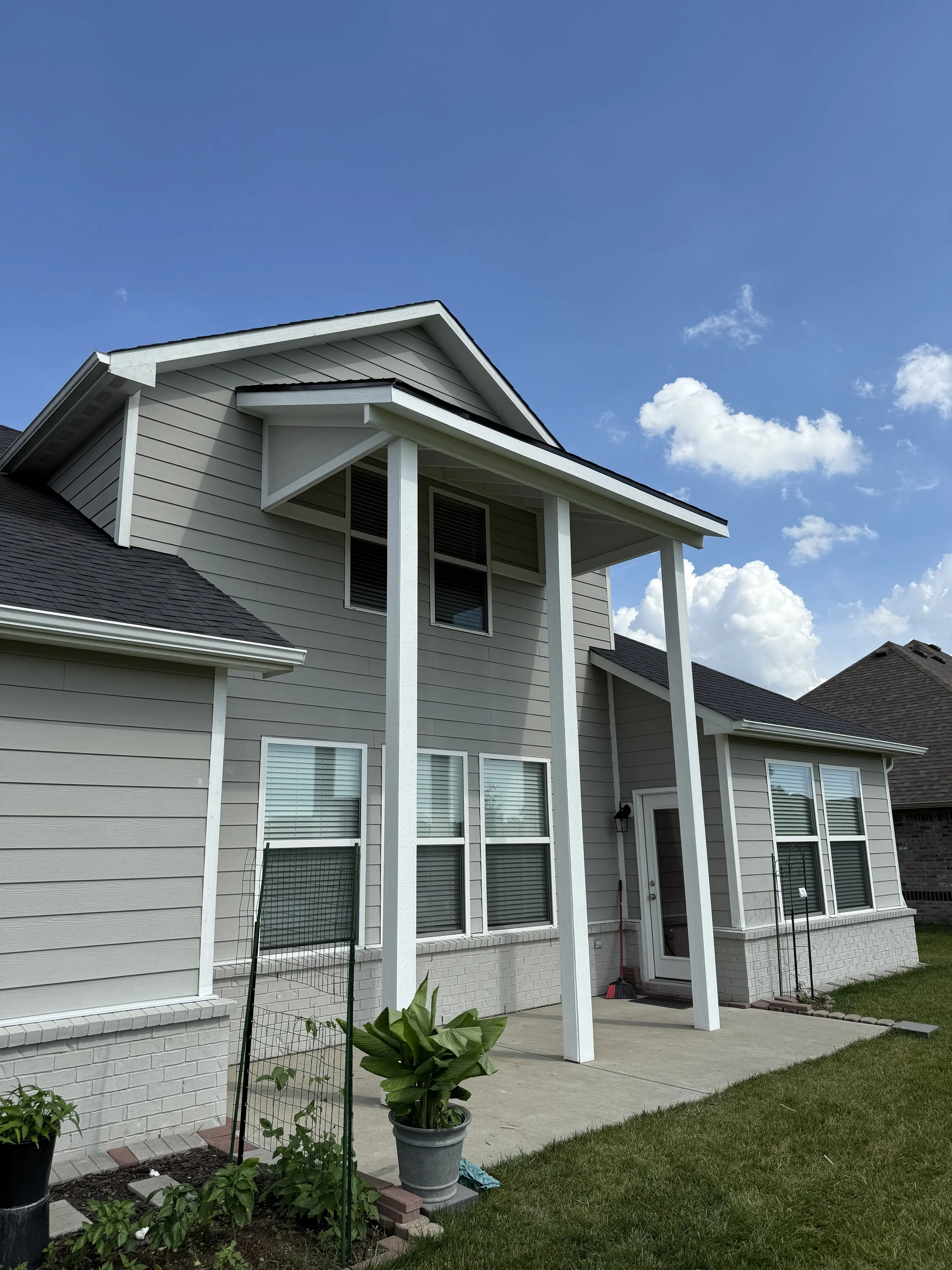 Back view of a two-story house with gray siding, white trim, and a small covered porch. There are four windows on the house, a door with a glass panel, and a garden with potted plants and a small garden plot in the yard. The sky is blue with a few cl