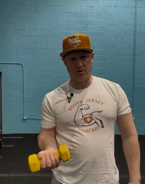 A man wearing a white South Jersey Armcare t-shirt and an orange baseball cap is holding a yellow dumbbell in a gym with a light blue brick wall background.