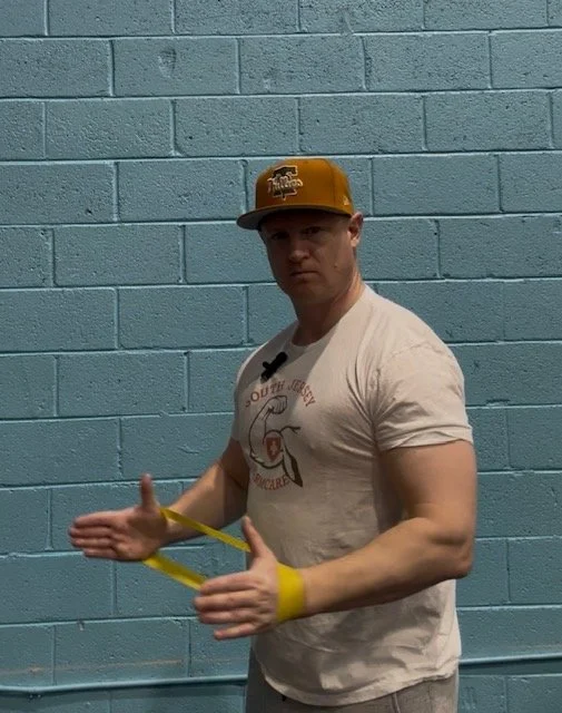 Young man in a beige t-shirt and yellow cap standing against a light blue cinder block wall, holding a yellow resistance band.