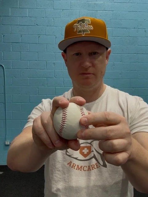 A person wearing a mustard yellow baseball cap and a white t-shirt with 'ARMCARE' written on it, holding a baseball in front of a blue brick wall.