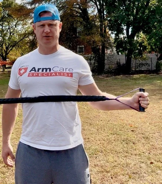 Man wearing a white t-shirt with ArmCare Specialist logo and a blue cap, holding a bow and arrow outdoors on a grassy field with trees in the background.