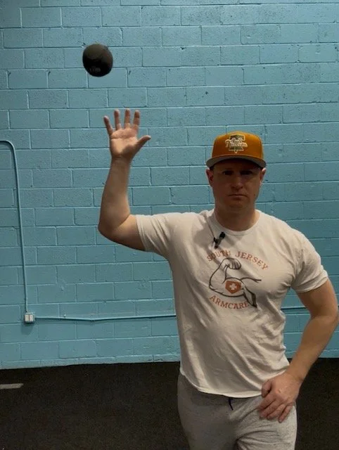 A young man in a T-shirt and cap is tossing a black ball into the air against a light blue brick wall.