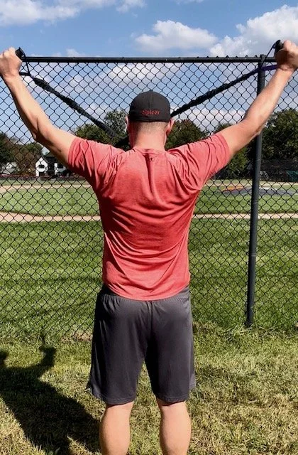 Person wearing a red shirt and black shorts stretching arms upwards while holding onto a chain-link fence with a grassy field and cloudy sky in the background.