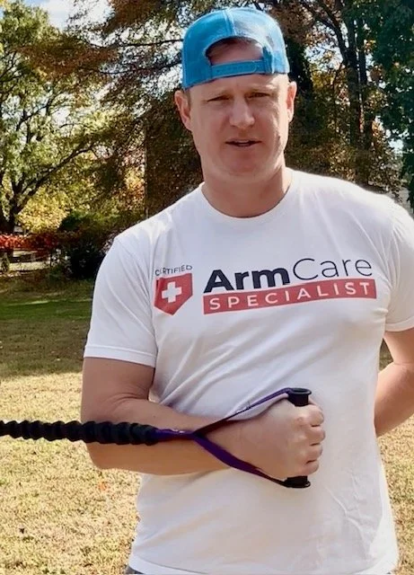 A man in a white T-shirt and a blue cap outdoors, holding a black object in his right hand. The T-shirt has a logo and text that reads 'Arm Care Specialist.'