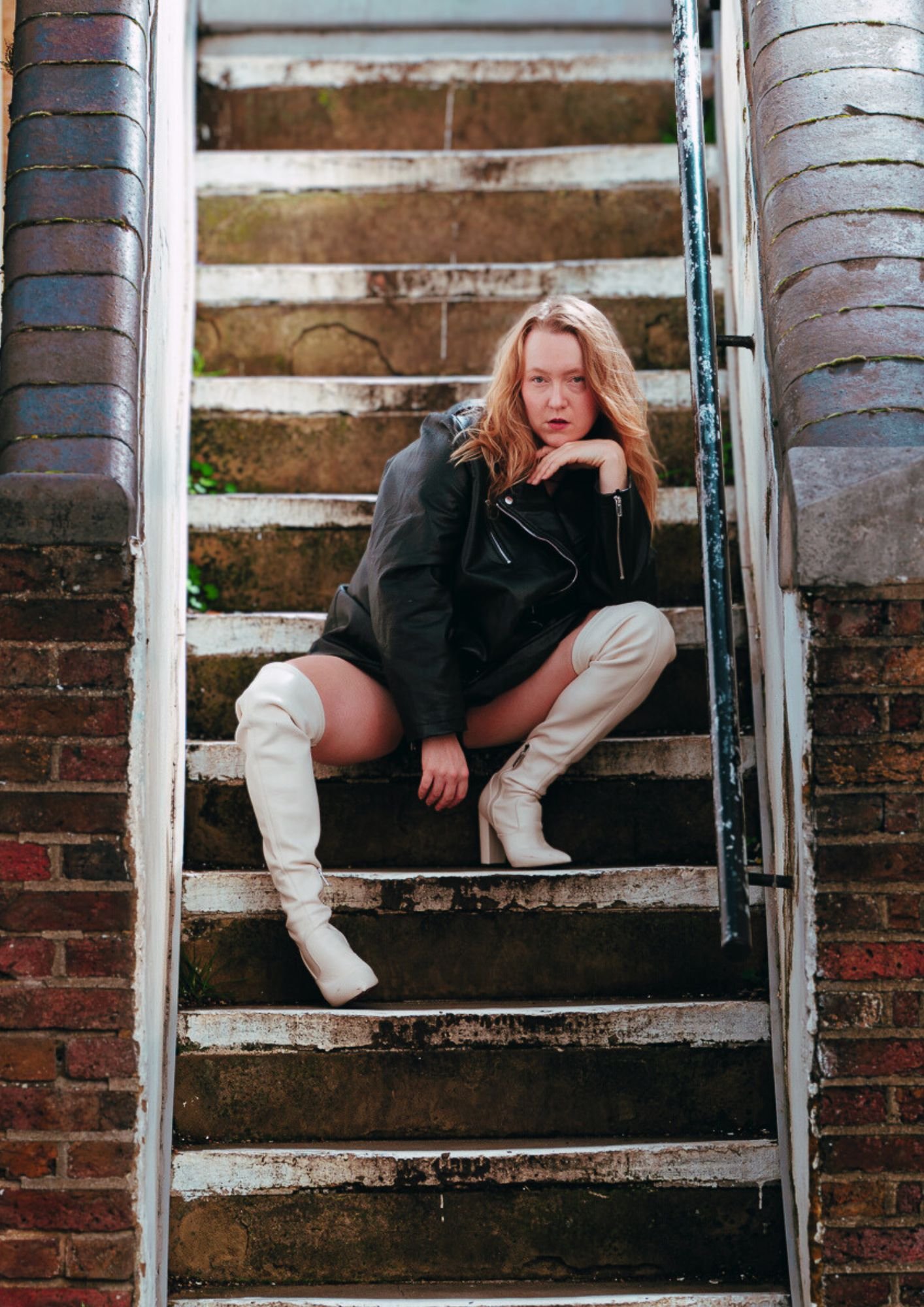 A woman with red hair kneels on outdoor brick stairs, wearing a black leather jacket and white thigh-high boots, looking directly at the camera with a confident expression.