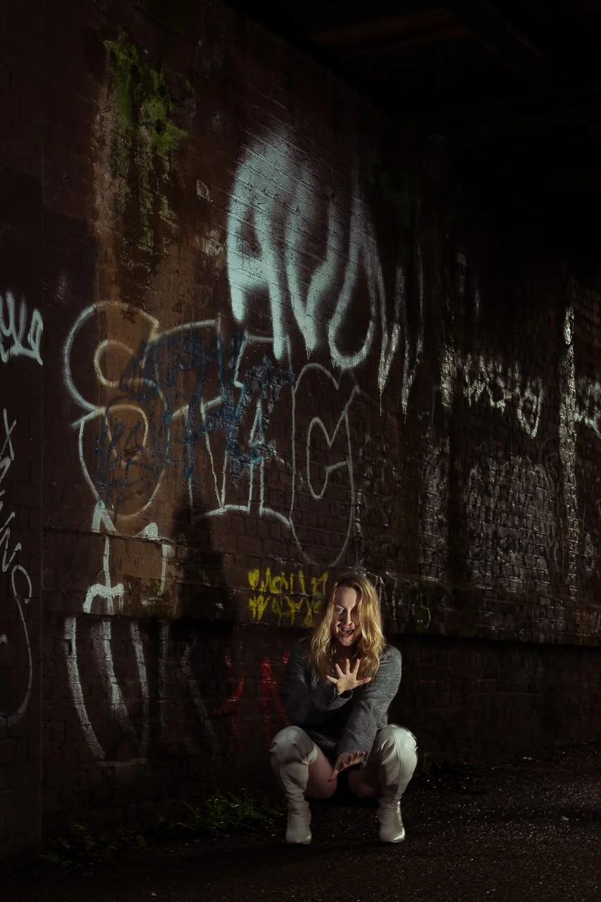 A woman crouching in front of a graffiti-covered brick wall at night, reaching out with one hand and smiling.