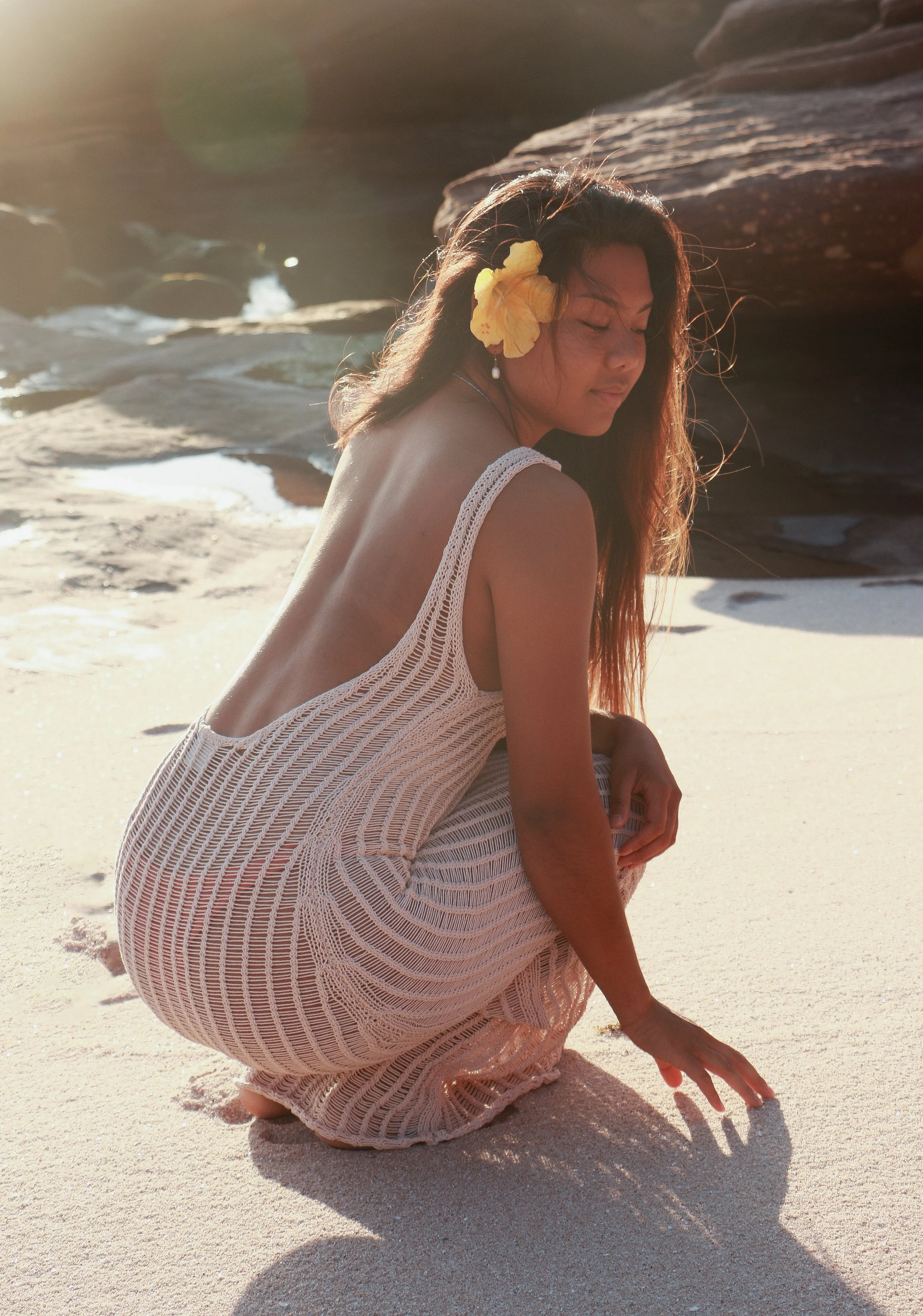 A woman with a yellow flower in her hair, wearing a white crochet dress, crouching on a sandy beach during sunset.